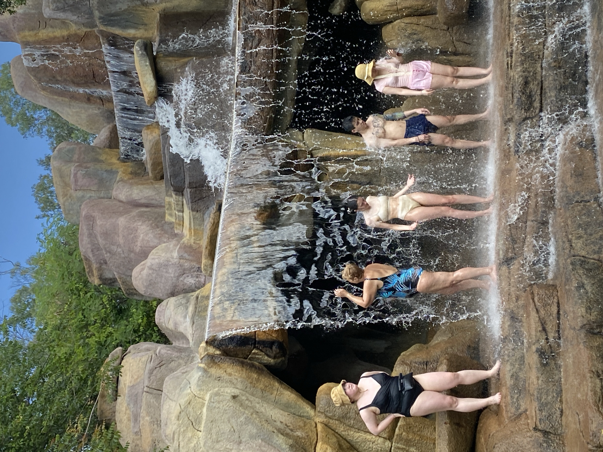 Five individuals are standing on tiered, wet rock formations in front of a man-made waterfall. The waterfall cascades over rocky ledges, creating a misty spray. The surrounding environment appears to be an outdoor recreational area, possibly a water park or resort, with lush green foliage and large rock structures. The sky is a clear blue.
From left to right, the individuals are:
1. A person wearing a black one-piece swimsuit and a tan bucket hat. They are facing left, with their hands on their hips.
2. A person wearing a blue and black patterned one-piece swimsuit. They are facing away from the camera, looking up at the waterfall, with their right arm raised.
3. A person wearing a beige two-piece bikini. They are facing the camera with their arms slightly extended to their sides.
4. A person, appearing to be male, wearing dark blue swim trunks. They are shirtless and facing the camera.
5. A person wearing a pink two-piece swimsuit and a tan bucket hat. They are facing the camera with their arms slightly bent.
The water is actively flowing, creating a dynamic and refreshing atmosphere. The rock formations are textured and appear to be designed to mimic a natural waterfall. The scene suggests a leisurely activity, likely involving cooling off in the water.