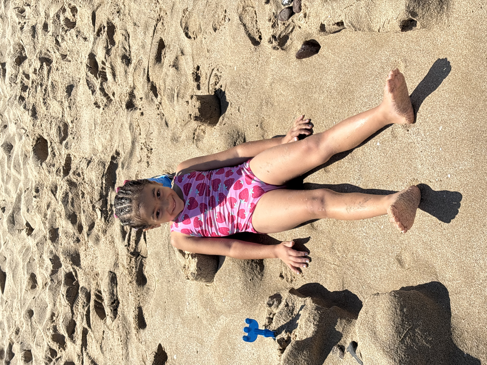 A young girl is lying on her back on a sandy beach. She is wearing a pink swimsuit with a strawberry pattern. Her hair is braided and tied with a pink ribbon. She is smiling and looking at the camera. A blue plastic toy rake is lying on the sand near her. The sand is wet in some areas, suggesting that the tide may have recently receded. There are footprints and small piles of sand around her, indicating that she may have been playing or building sandcastles. The sun is shining, casting shadows on the sand, which suggests it is daytime. The scene appears to be a sunny day at the beach in Primorsko, Bulgaria.