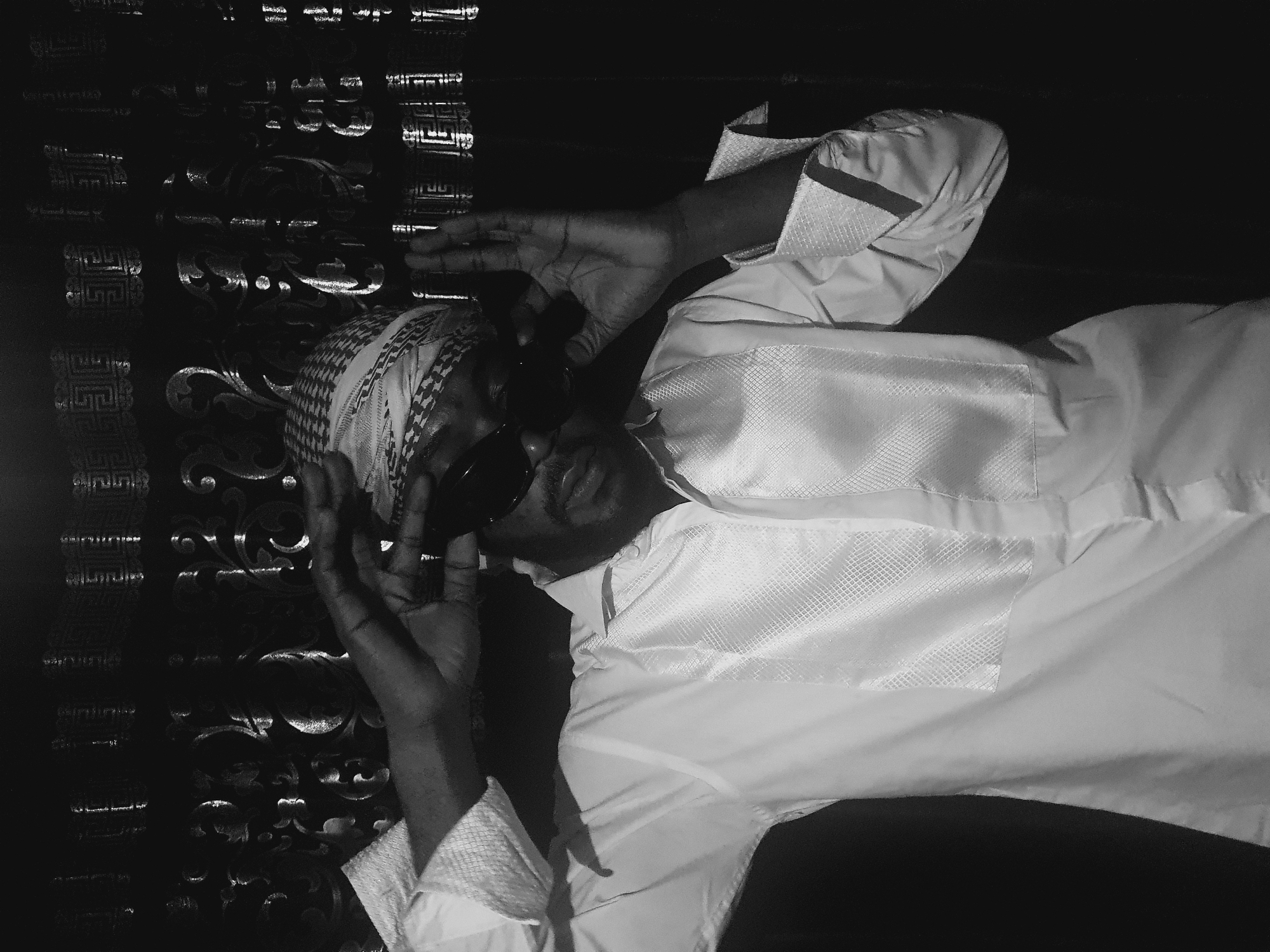 A man wearing a ghutrah and sunglasses is pictured in front of a decorated surface. The man is wearing a light-colored robe and holding his hands near his face. The photograph is in black and white. There is no visible indication of Lokoja, Nigeria.