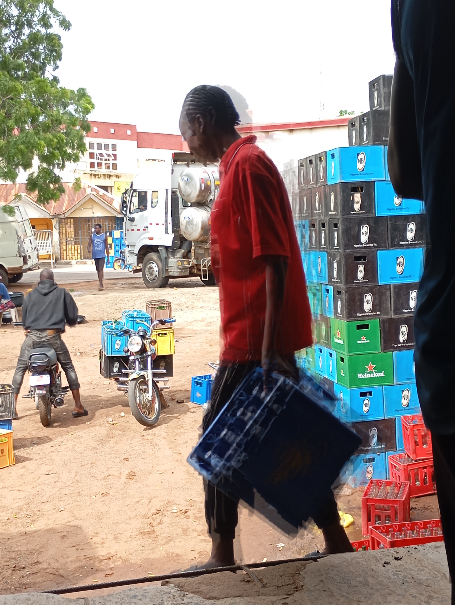 In the city of Shendam, Nigeria, on a dirt-covered ground under a bright sky, a man in a red shirt is blurred by motion as he carries a stack of blue plastic crates. Behind him, a large pile of black and blue crates, some with "NB Nigerian Breweries Plc" labeling, are stacked high. To the left, a man in a black hoodie and jeans sits on a motorcycle, which is loaded with blue and yellow crates. A white truck with large cylindrical tanks is parked in the middle ground, and in the background, buildings with red trim are visible. Other individuals and vehicles are present, contributing to a busy, commercial atmosphere.