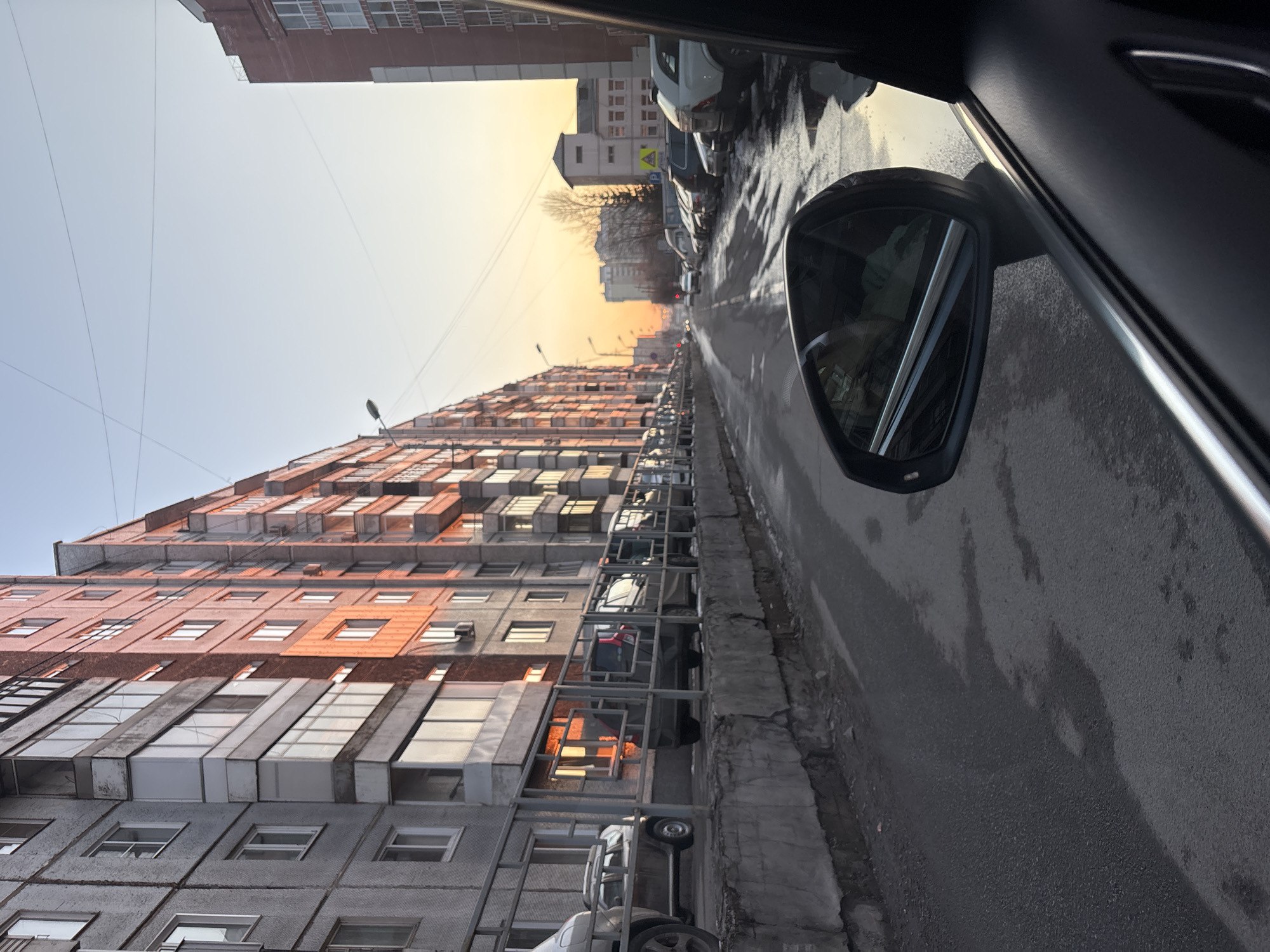 The photograph, taken from inside a vehicle, captures a street scene in Krasnoyarsk, Russia. The view looks down a wet, asphalt road lined with parked cars. On the left, a tall, multi-story residential building dominates the scene, displaying a mix of gray and orange facades. The sky is pale, suggesting either dawn or dusk. The side mirror of the car is visible on the right, reflecting the street and surrounding environment. There are no visible people.
