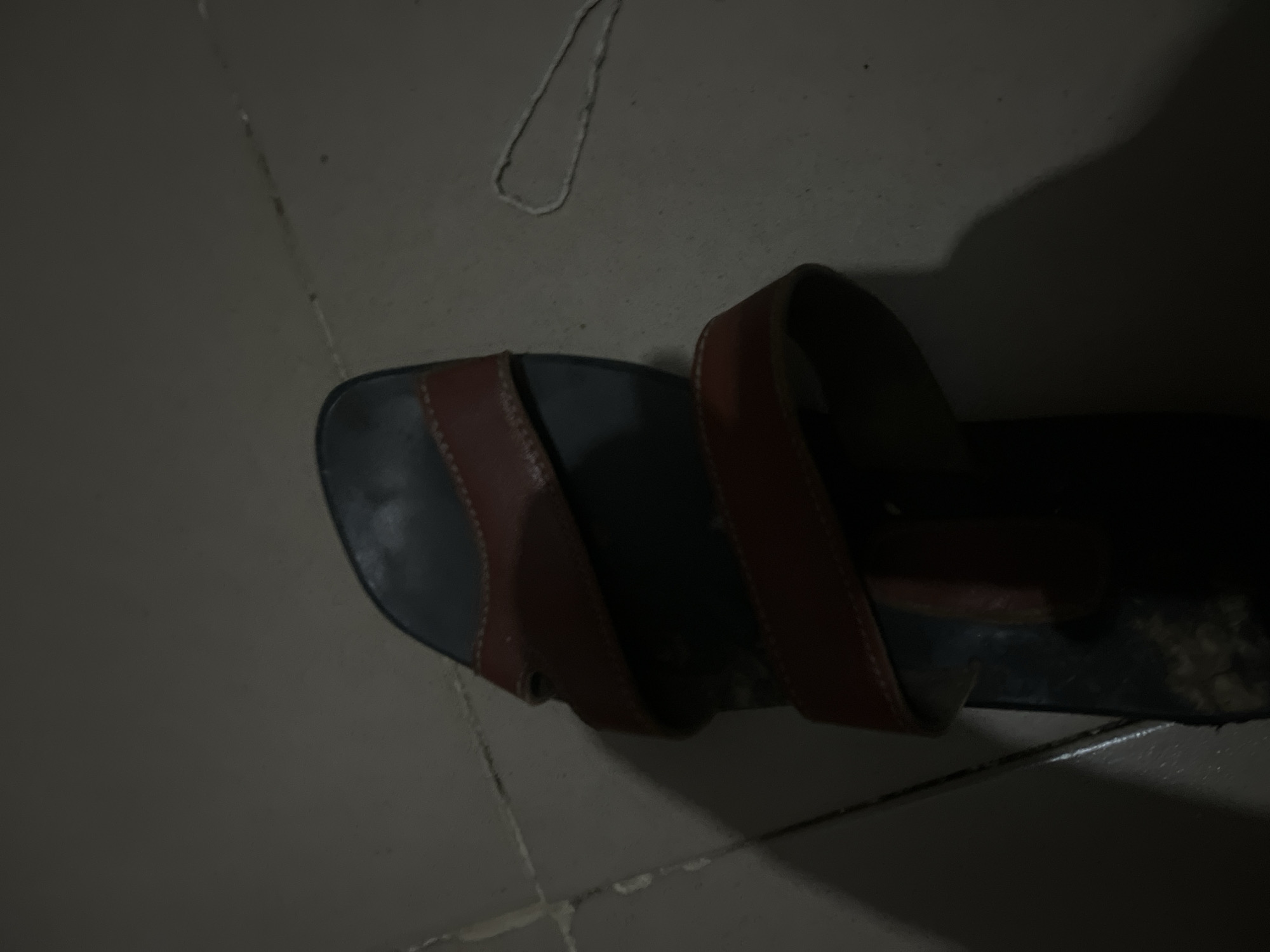 An overhead, close-up shot shows a single, brown and black sandal on a tiled floor. The sandal appears to be made of leather and has two straps that go across the foot. The sole of the sandal is dark, possibly black, and the straps are a reddish-brown color. The sandal is oriented vertically in the image, with the toe pointing towards the bottom and the heel towards the top. The tiled floor is a neutral color, with visible grout lines. There is a shadow cast by the sandal on the right side of the image. A thin white string or thread is visible on the floor to the right of the sandal. The lighting in the image is dim, suggesting it was taken in low-light conditions. No people are visible in the image. The location is an indoor setting, possibly a room in Jalingo, Nigeria, given the context.
