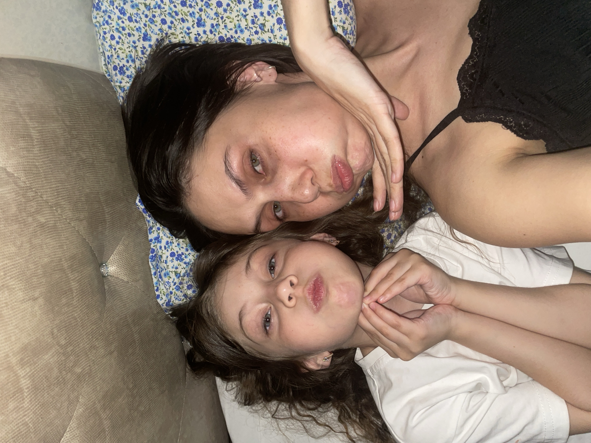This indoor photograph, taken in Izhevsk, Russia, features two females: an adult woman and a young girl, likely mother and daughter, lying down together. They are positioned with their heads close, both looking directly at the camera.
The adult woman, with dark hair, lies on a pillow with a blue and white floral pattern. She is wearing a black top with lace detailing. Her face shows a playful expression with pursed lips, often called a "fish face," and she has green eyes. One of her hands is raised and positioned near her face.
Below her, the young girl, also with dark, curly hair, is wearing a white t-shirt. She mimics the woman's expression with pursed lips and has bluish-gray eyes. Her hands are brought up to her chin, framing her face in a similar pose.
They appear to be on a light brown or beige sofa or bed, which has a textured, possibly quilted design with a decorative button or stud visible. The lighting is soft, suggesting an indoor setting, possibly in the evening. The overall mood is playful and affectionate, capturing a bonding moment between the two. There is no visible text in the image.