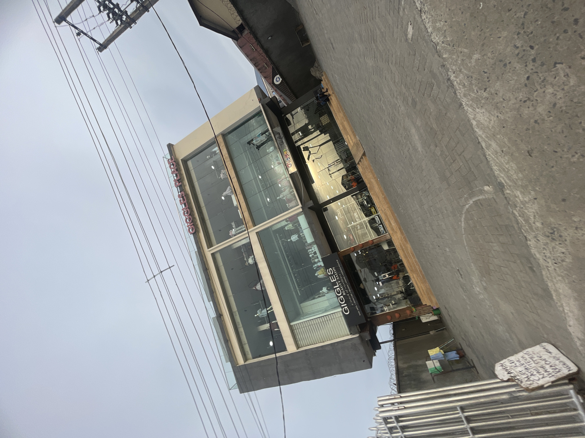 The image depicts a daytime street scene in Ado, Nigeria, featuring a multi-story commercial building under an overcast sky. The modern building, likely a salon or retail store, has a prominent glass facade across its upper floors, reflecting parts of the surroundings. A large black sign on its lower facade reads "GIGGLES BEAUTY SALON" with the slogan "a touch of excellence" and a list of services including "NAIL ARTS PEDICURE BRAIDIN". Below this, another line reads "Giggles Tabs". Above the main sign, the word "GIGGLES" is displayed in red letters on the building's upper section.
In the upper portion of the frame, a utility pole with numerous overhead power lines stretches across the dull, grey sky, creating a dense network of wires. The ground level, visible on the left, consists of both grey interlocking paving stones and rougher concrete. On the lower right, a stack of metal railings or bars is present.
A distinctive detail is a handwritten white sign placed on the ground, advertising various job positions. The legible text on this sign includes "CASHIER", "SALES REP", "HAIR STYLIST", "NAIL TECH", "BRAIDER", and "NEEDS WAGES". Other partial words such as "NAUTECH", "BRAGEP", and "ALES REP" are also visible. No direct human activity is captured, but the presence of the salon and the job recruitment sign suggests an active urban commercial environment during the day, indicated by the ambient, diffused light and lack of strong shadows.