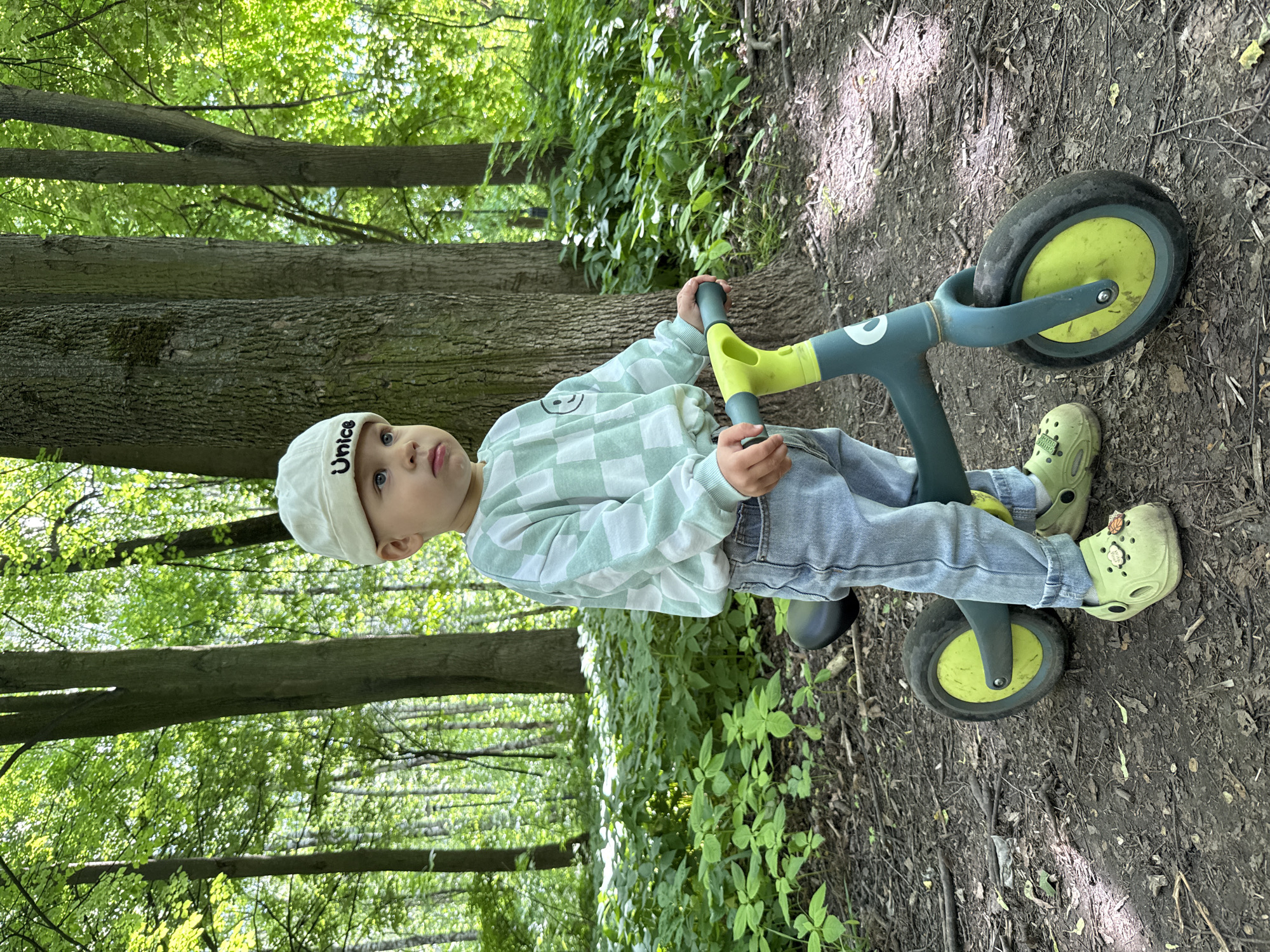 A young child, likely a toddler or preschooler, is captured in a daylight outdoor setting, possibly a park or wooded area in Rusanovo, Russia. The child is seen sitting on a green and yellow balance bike, holding the handlebars, and looking directly towards the camera with a calm expression. They are wearing a white baseball cap with the word "Unice" printed on it, a light green and white checkered long-sleeved shirt featuring a small smiley face, light blue jeans, and light green Crocs.
The setting is characterized by a dirt path strewn with leaves and small branches, indicating a natural environment. Tall, deciduous trees with vibrant green foliage line the background and sides of the path, suggesting a spring or summer day. Bright sunlight filters through the tree canopy, creating dappled light on the ground and illuminating the scene. The image portrays a serene moment of a child paused during outdoor activity.