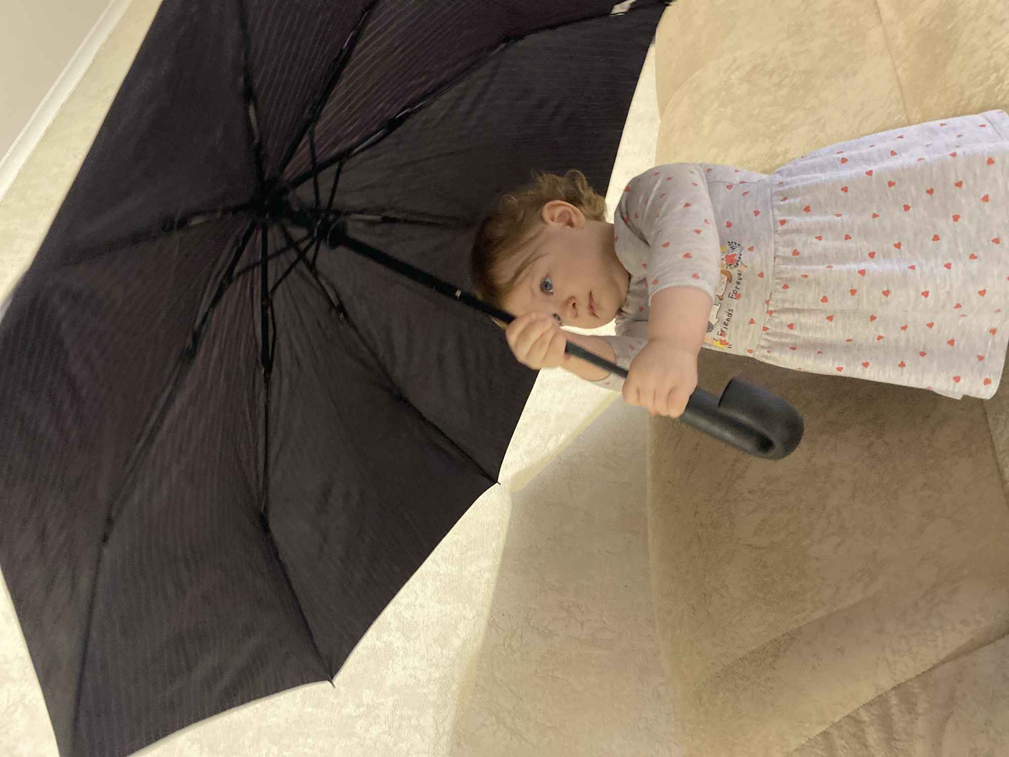 A toddler lies on a beige couch, holding a closed, black umbrella. The child is facing the viewer, with fair skin, brown hair, and blue eyes. The toddler wears a light gray dress patterned with small red hearts and an image of cartoon figures. The umbrella's canopy fills the left side of the frame, while the couch occupies the right side and background. The location appears to be indoors.