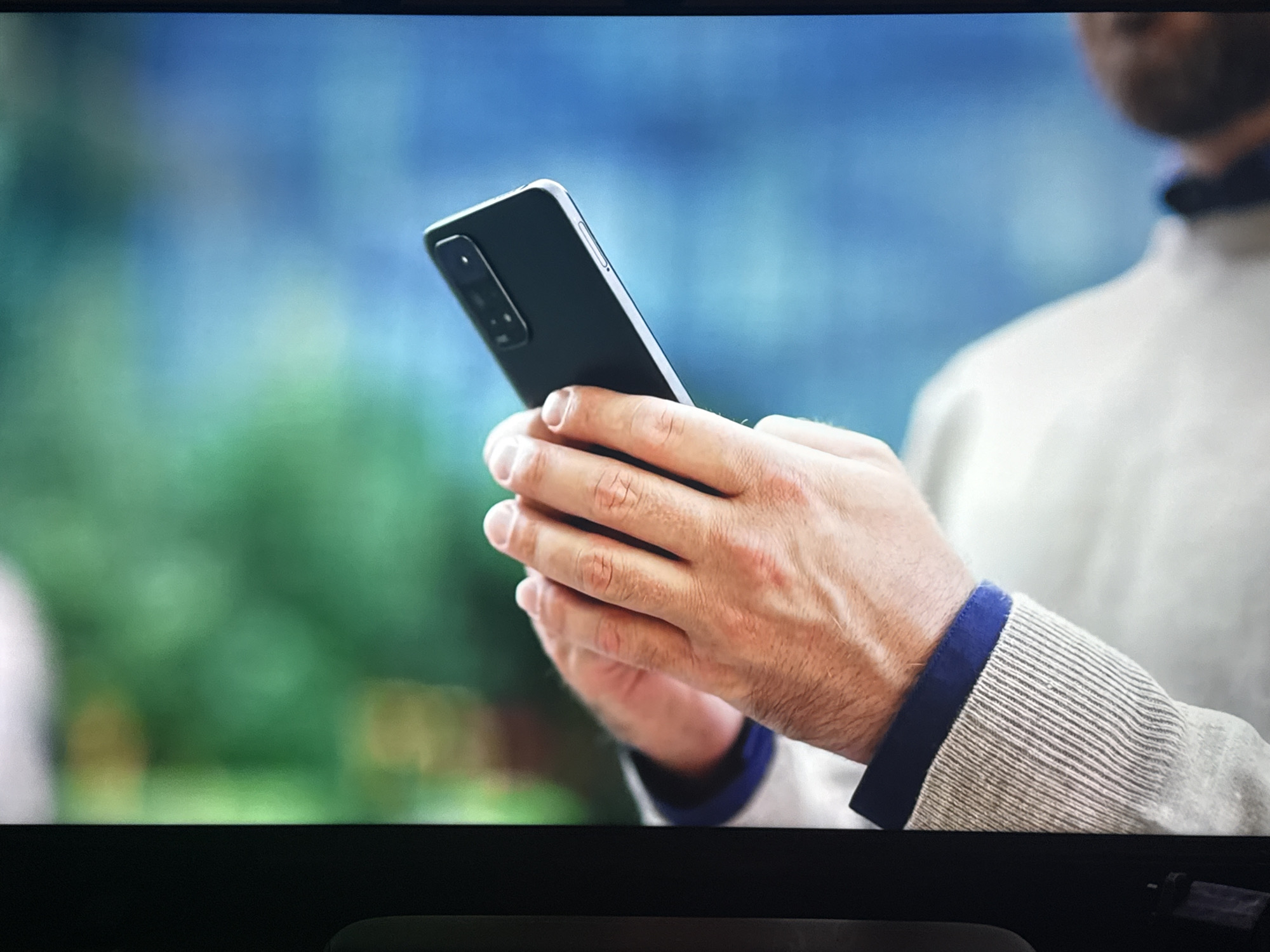 A person, partially visible from the shoulder up, is holding a black smartphone in their hands. The person is wearing a light-colored sweater with blue trim on the cuffs. The background is blurred, showing a mix of green foliage and a blue sky, suggesting an outdoor setting. The image is captured on a screen, likely a television or monitor, as indicated by the edges of the screen visible at the top and bottom of the frame. The location is identified as Cheltenham, United Kingdom.