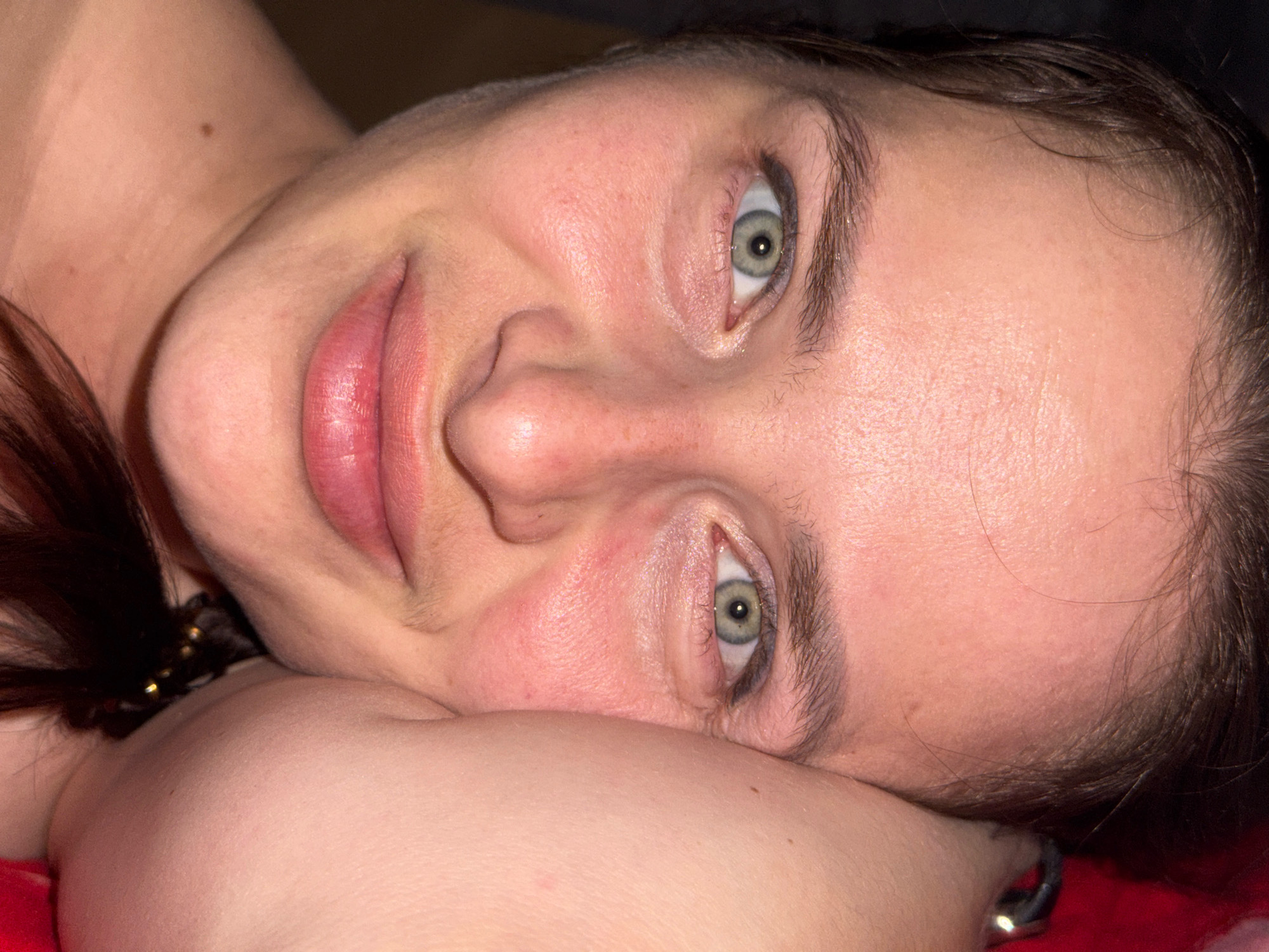A close-up, low-angle shot shows a young woman lying down, with her head resting on her arm. Her eyes are open and looking directly at the camera. Her hair is dark and cascades down her left side. The woman is wearing a necklace with gold beads. The background is dark, with some red fabric visible in the lower right corner. There is no indication of the location being in Rusalevka, Russia.