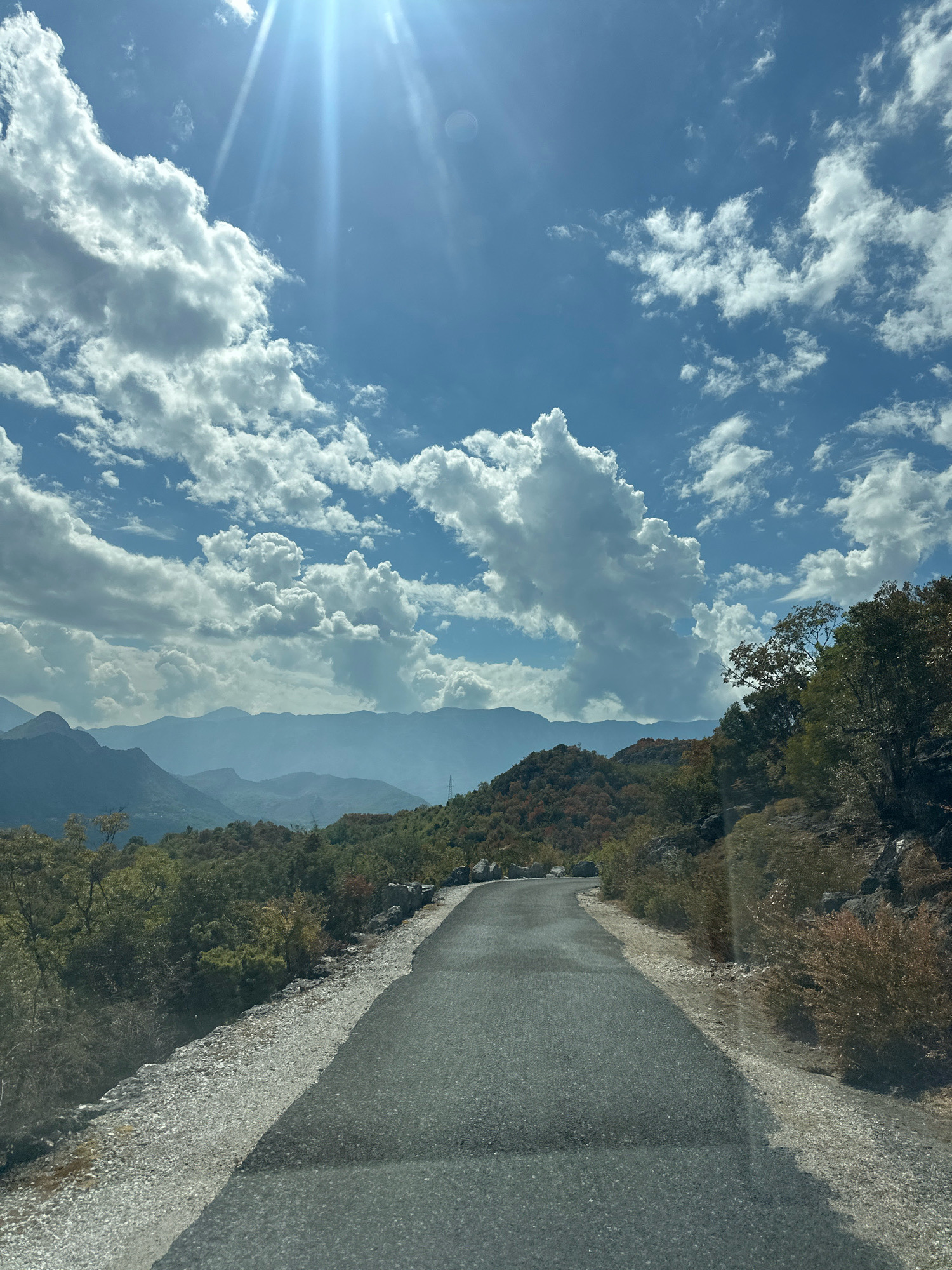 The image shows a paved road winding through a mountainous landscape under a partly cloudy sky. The sun is shining brightly, casting lens flares and rays through the clouds. The road is in the foreground, leading towards a distant range of hazy blue mountains. The surrounding terrain is covered in dense vegetation, with trees and shrubs on the hillsides. The sky is a vibrant blue, dotted with large, fluffy white clouds, some of which are illuminated by the sun. On the right side of the road, there is a rocky slope covered with dry brush and scattered trees. The overall impression is of a scenic, rural environment. The location is identified as Kruševica, Montenegro. There are no people or specific objects that would indicate any action or interaction.