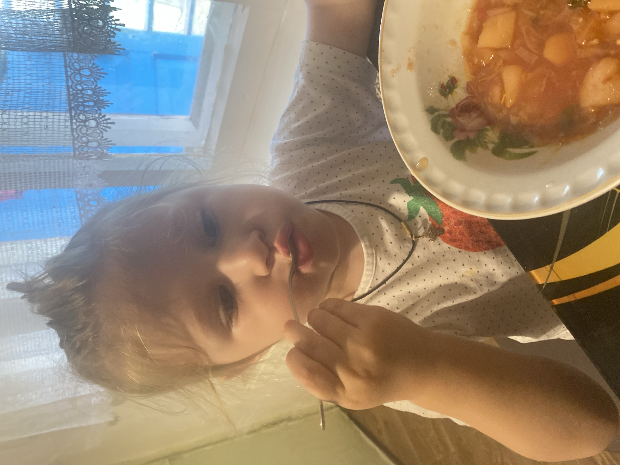 A young child with blonde hair is eating from a plate of food. The child is wearing a white t-shirt with a pattern of small black dots and a strawberry graphic. A necklace is visible around the child's neck. The child is holding a spoon and bringing it to their mouth, with the spoon containing some of the food. The plate is white with a floral pattern and a gold rim. The food appears to be a stew or soup with chunks of vegetables and possibly meat.
The scene is indoors, with a window visible in the background. A lace curtain hangs in front of the window, and through the window, a blue sky is visible. The lighting suggests it is daytime. The child's facial expression is neutral, possibly focused on eating. The overall setting appears to be a domestic environment, possibly a dining area. The provided location context is Маринопіль, Ukraine.