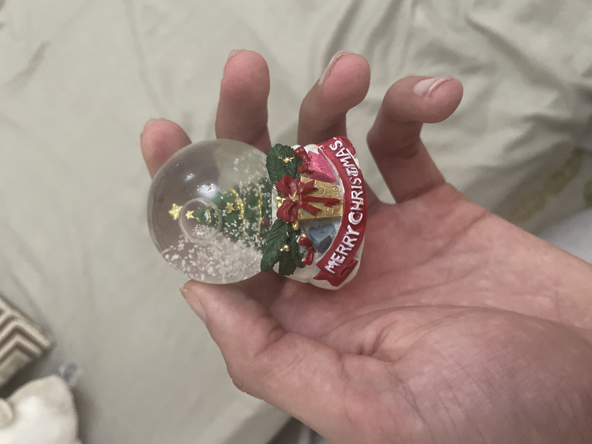 A person's hand is centrally positioned, holding a small Christmas snow globe. The snow globe features a clear spherical glass top enclosing a miniature green Christmas tree adorned with a golden star and small decorations, surrounded by white artificial snow flakes. The snow globe rests on a decorative base, which is white with green garland, miniature wrapped presents in gold, pink, and blue/grey hues, and a red banner inscribed with "MERRY CHRISTMAS" in white capital letters. The hand is holding the base of the globe between the thumb and fingers. The background consists of a light-colored, possibly off-white or light green, rumpled fabric surface, likely bedding. In the blurred upper left background, indistinct light-colored and patterned objects are visible. The specific location in the city of Ihtiman, Bulgaria, cannot be determined from the visual information provided in the image.