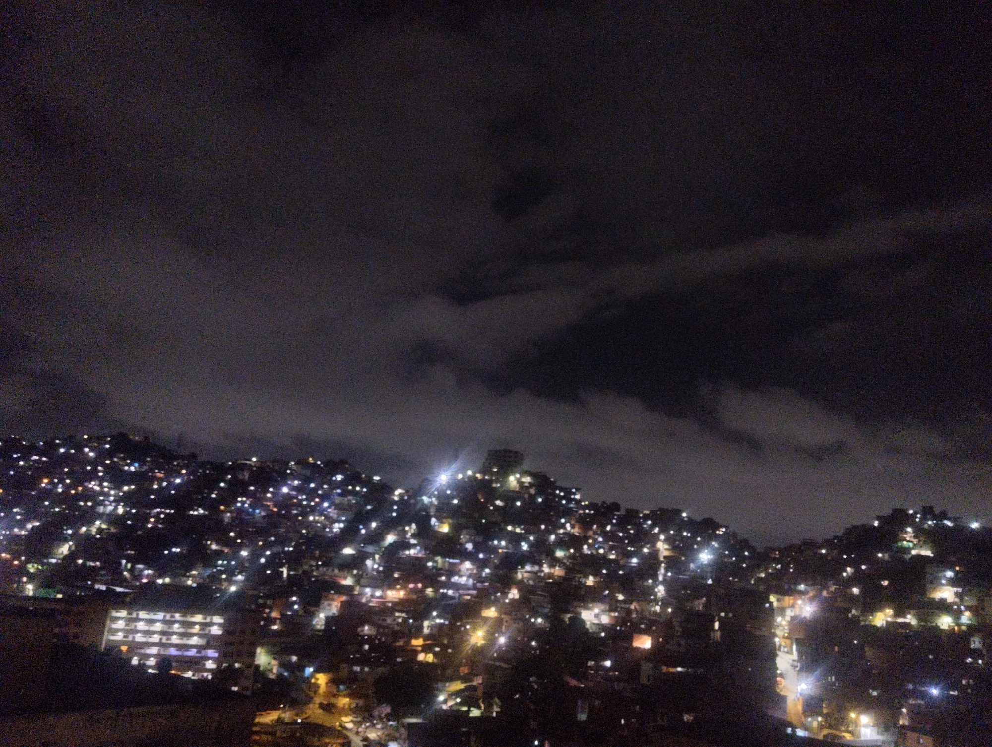 The image shows a nighttime view of a densely populated area, likely a hillside favela, within the city of Caracas, Venezuela. Thousands of lights from dwellings are scattered across the hills, indicating residential occupation. There is a dark, cloudy night sky. The lower foreground exhibits a darker area, presumably lower-lying structures and streets. A prominent, brighter building with multiple illuminated windows is visible at the bottom left of the image.