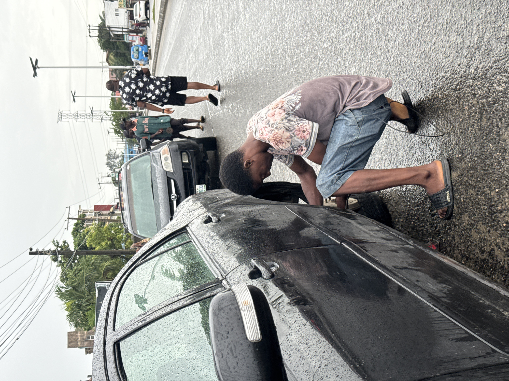 A man wearing a floral shirt and blue shorts is bending down next to a black car, apparently changing a tire, on a wet street in Ado, Nigeria. The car has a partially visible license plate: "KJA6JO." Three other people are in the background, walking on the street past a black SUV. The street appears wet, suggesting recent rain. Power lines and vegetation are visible in the background, and there are also buildings and other vehicles further down the street.