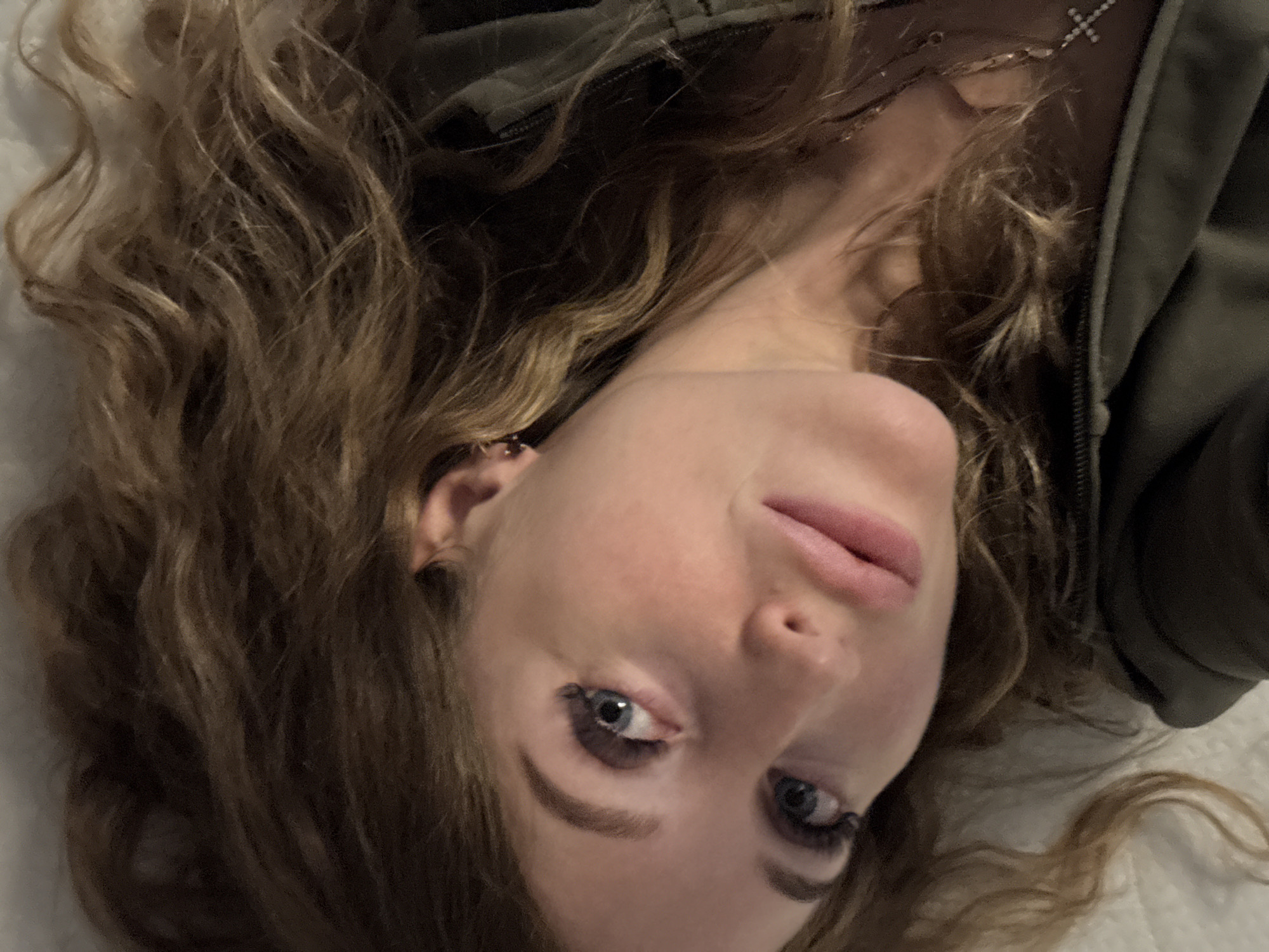 A woman is lying on her back. She has light skin, light brown, curly hair, and blue eyes. She is wearing a green jacket and a necklace with a cross. The background appears to be a white surface. There is no indication of the location being Chaikovsky, Russia.