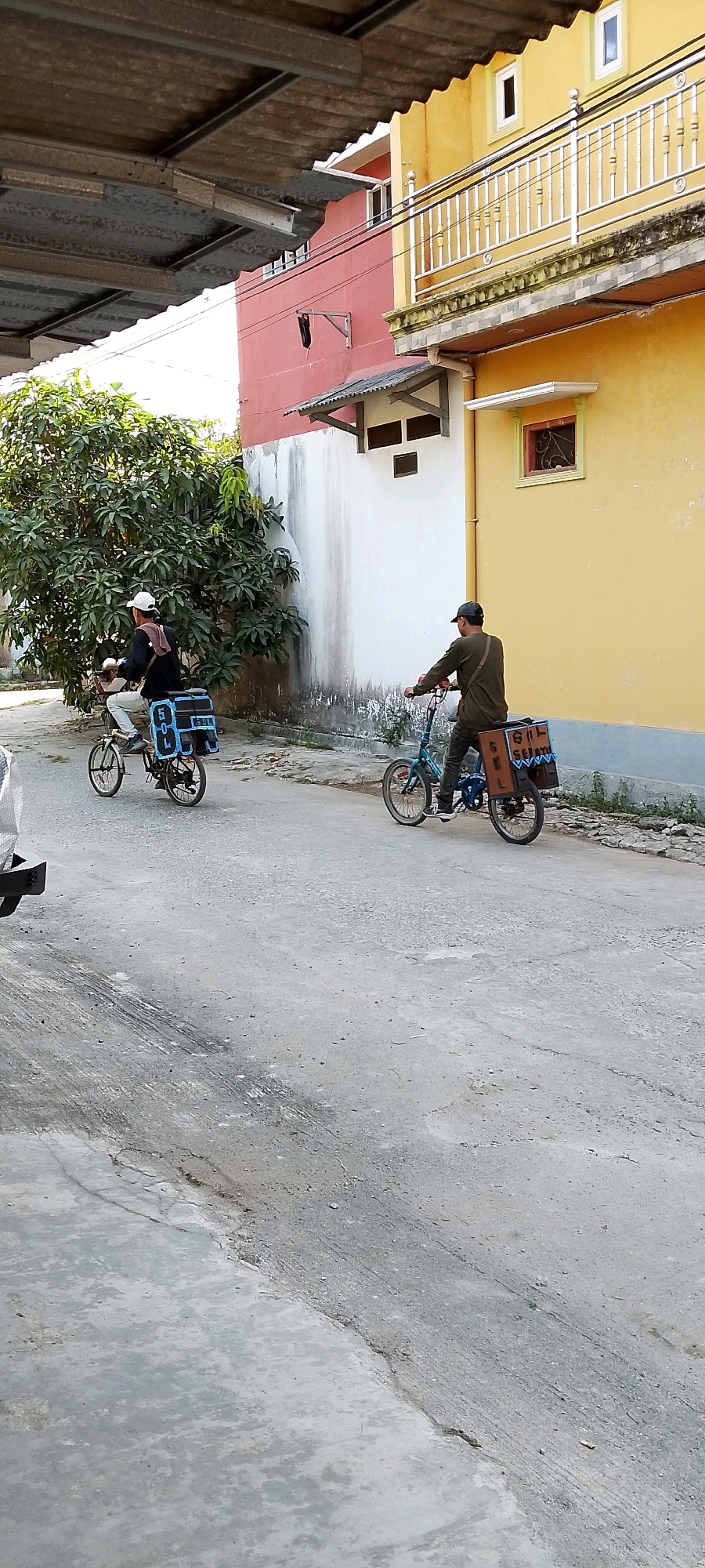 In Satriamekar, Indonesia, on a paved street, two individuals are riding bicycles equipped with cargo boxes on the rear. The bicycle on the left is being ridden by a person wearing a black shirt, white hat, and glasses, with a dark bag over their shoulder. The bicycle on the right is ridden by a person in a dark green long-sleeved shirt and a dark cap, also with a dark bag. Both bicycles have blue and brown cargo boxes. The background features a yellow building with a balcony and a pink building adjacent to it, separated by a white wall. A large leafy tree is visible to the left of the pink building. The ground is a mix of worn concrete and asphalt.