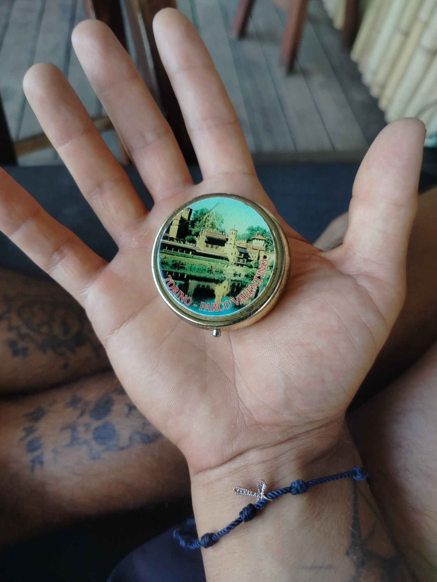 A person holds a round, gold-rimmed souvenir in their open palm. The souvenir depicts a building reflected in water, with the text "TORINO - PARCO VALENTINO" encircling the image. The person has a blue braided bracelet with a small silver cross charm on their wrist. Tattoos are visible on the person's thigh. The location is Torino, at the Parco Valentino.
