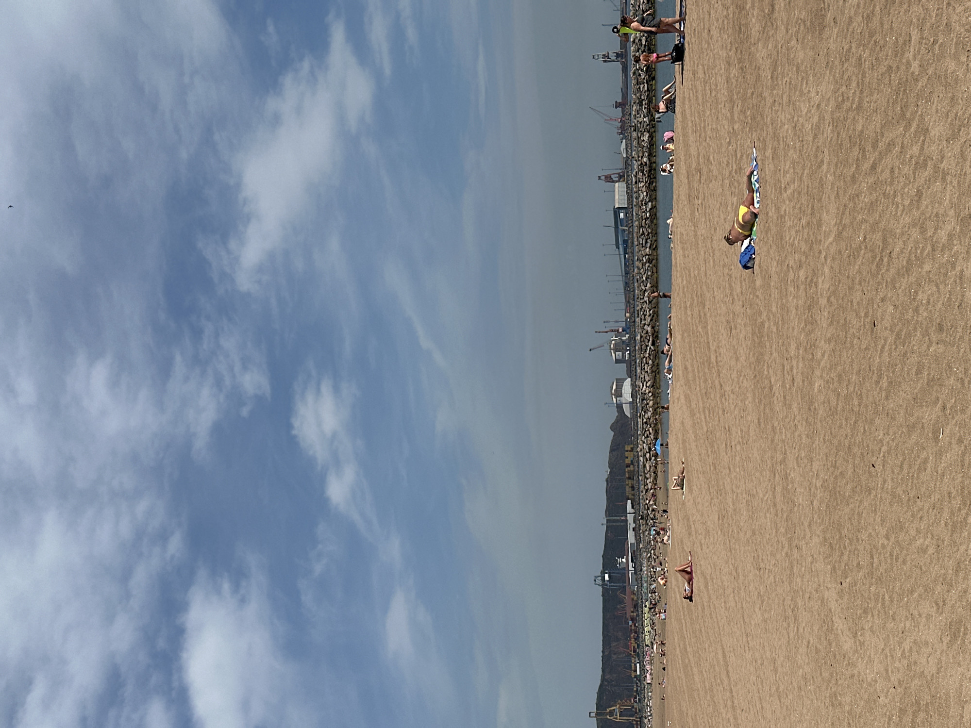 The image captures a beach scene in Gijón, Spain, juxtaposing the natural landscape with industrial elements. A wide sandy beach occupies the right side of the frame, dotted with people sunbathing and enjoying the coast. A woman in a yellow bikini lies on a towel on the beach. The left side is dominated by a blue sky filled with scattered clouds. In the middle ground, a seawall separates the beach from the harbor area, where various industrial structures like cranes and buildings are visible. The water is a darker shade, creating a contrast with the sky. People are gathered along the seawall and on the beach, some appearing to walk or stand near the water's edge. The scene conveys a sense of leisure activities occurring in close proximity to an active port.