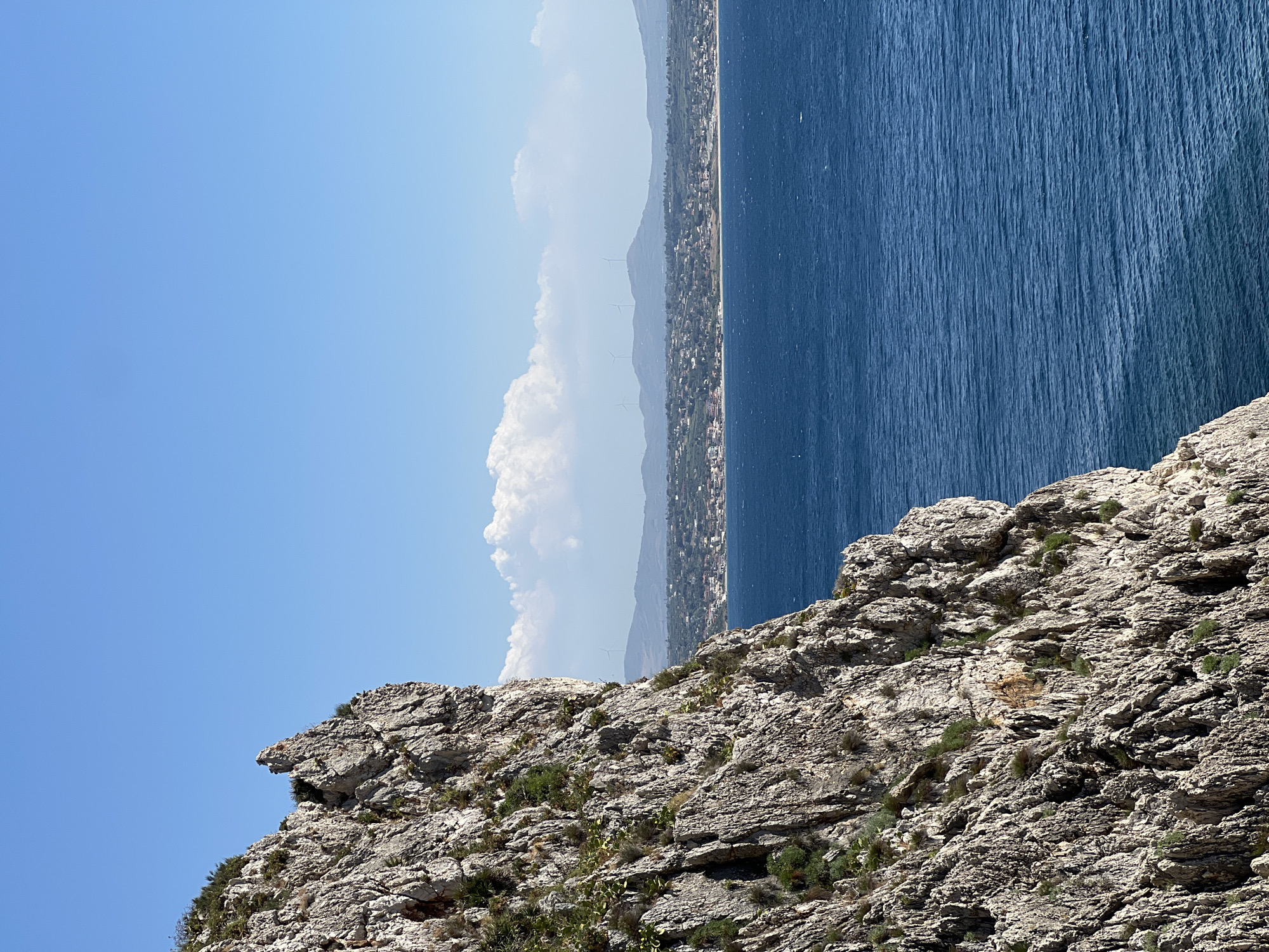 This image depicts a rocky, coastal landscape on a clear, sunny day. The foreground is dominated by rugged, grey rocks covered in sparse green vegetation, suggesting a dry, Mediterranean climate. Beyond the rocks, a vast expanse of deep blue sea stretches out, its surface rippled by gentle waves. In the middle ground, a coastal town is visible, nestled along a strip of land between the sea and distant, hazy mountains. The architecture of the town appears densely packed, with red-tiled roofs characteristic of the region. Further in the distance, several wind turbines can be seen on the hills, indicating a location that utilizes renewable energy. The sky is a brilliant, cloudless blue, with a few wispy white clouds near the horizon. The lighting suggests it is daytime, likely late morning or early afternoon, given the brightness and the clear shadows cast by the rocks. The overall impression is one of a serene, picturesque coastal scene in Terrasini, Italy, likely during the summer months. There is no visible text in the image.