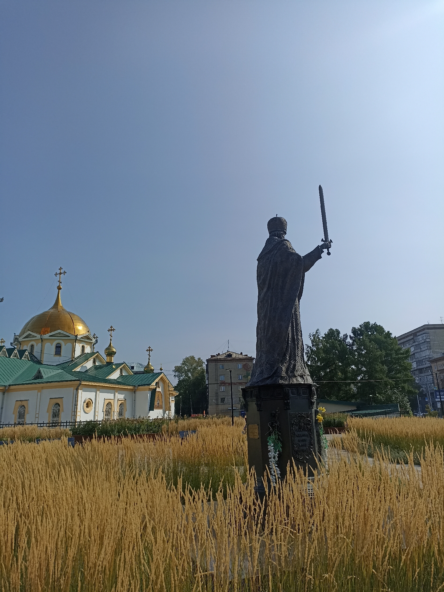 The image shows a statue of a robed figure wearing a crown and holding a sword aloft. The statue is positioned in front of a white building with a golden dome and green roof, characteristic of Orthodox churches. This building is identified as the Ascension Cathedral in Novosibirsk, Russia. The foreground is filled with tall, dry grass or reeds. In the background, there are trees and modern buildings, indicating an urban setting. The sky is clear and blue.