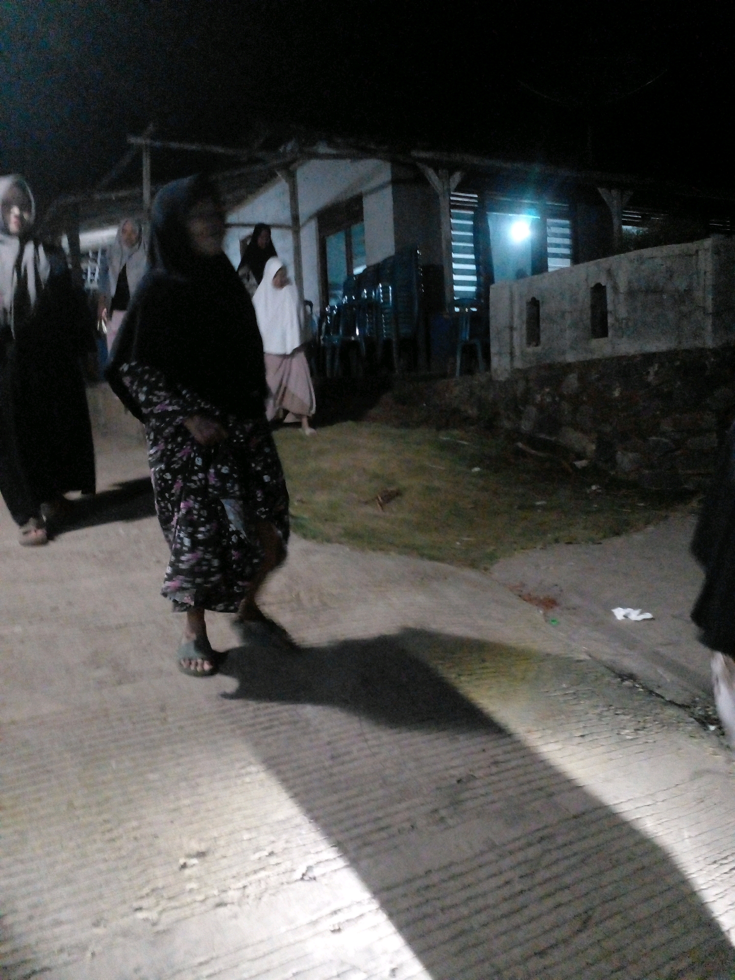 In the city of Pungangan, Indonesia, at night, several individuals are visible on a concrete pathway. One person in the foreground is wearing a dark headscarf and a long, dark skirt with a floral pattern. They are also wearing sandals and are casting a shadow on the ground. To the left, another person is seen wearing a light-colored headscarf and a dark garment. Further in the background, other figures are present, including one wearing a white garment. The environment appears to be an outdoor area with a building structure behind the individuals. There is grass and some debris on the ground. A bright light source is visible in the background, illuminating part of the scene. The overall lighting suggests it is dark, with artificial light sources contributing to the visibility.
