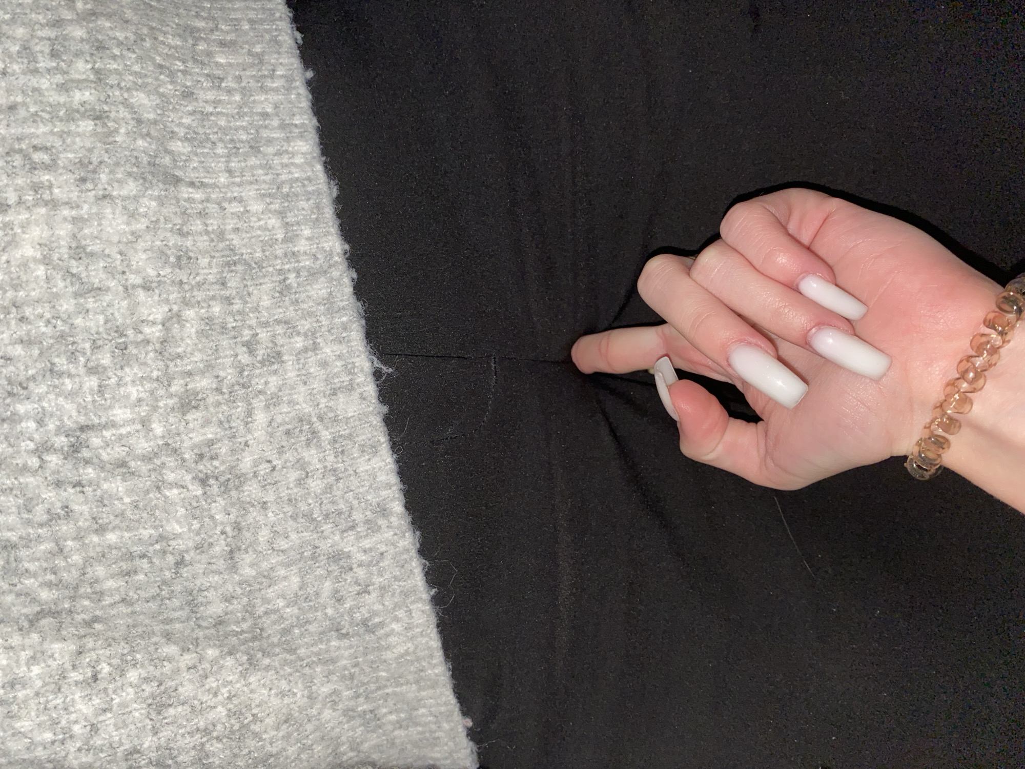 The image displays a close-up view of a person's hand on a dark fabric surface, positioned next to a lighter, textured fabric. The frame is horizontally divided by these two textile materials. On the left, a light gray, fuzzy or knitted fabric with a visible texture fills about a third of the width. The remaining portion of the frame on the right is occupied by a smooth, dark black fabric.
A pale-skinned hand, likely female, extends from the right side of the frame, with its index finger pressing into the black fabric, creating a subtle crease. The hand features long, uniformly white, coffin-shaped artificial nails on all visible fingers (thumb, index, middle, ring, and pinky). A translucent, light brown or amber-colored coiled hair tie is worn as a bracelet on the wrist.
The lighting suggests an indoor setting. There are no other visible subjects, activities, specific location cues, or text. The focus is on the hand, its accessories, and the contrasting fabric textures.