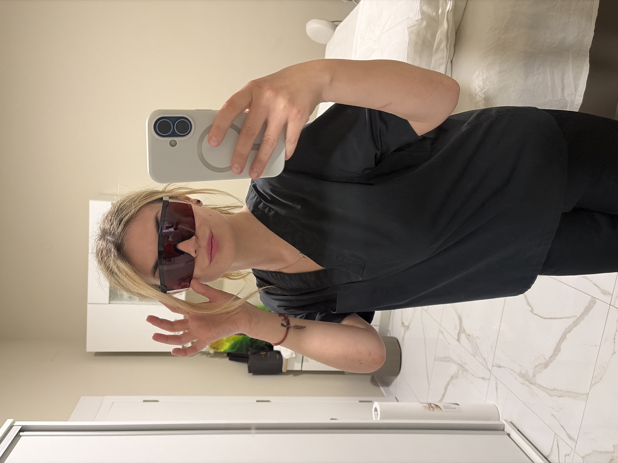 A mirror selfie taken indoors in Budva, Montenegro, shows a woman with blonde hair wearing dark-lensed, black-framed sunglasses and a dark-colored top, possibly a uniform or scrubs. She holds a light-colored smartphone with a MagSafe-style case and two prominent camera lenses, angled to capture her reflection. Her lips are adorned with light pink lipstick. On her right wrist, she wears a red string bracelet with a small, light-colored charm, and a small dark tattoo is visible on her inner forearm.
The background reveals a contemporary room with a minimalist aesthetic. To the left (from the viewer's perspective), a bed with white bedding is partially visible. To the right, a white cabinet or dresser is seen, with a dark object resting on a shelf above it. The floor features white tiles with prominent gray marble-like veining. The walls are a light cream or beige color. The room appears brightly lit, suggesting either natural daylight or ample artificial lighting. No visible text is present in the image.