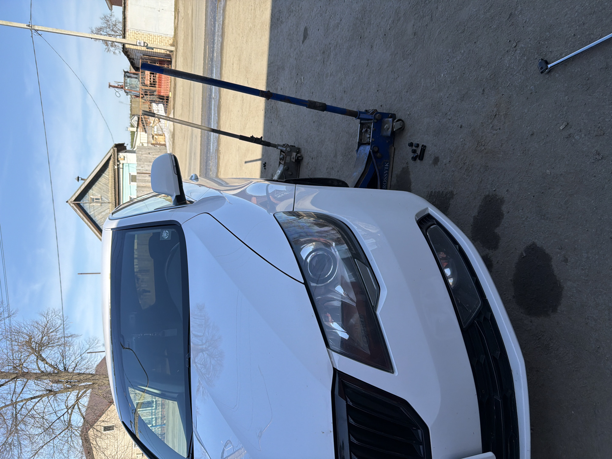The image shows a white car undergoing maintenance. The car is jacked up using a blue hydraulic jack, with a smaller, similar jack also present. Several lug nuts are scattered near the jack. The car is parked on a paved surface with some dark fluid stains.
In the background, there are residential buildings typical of a small town or village. A blue sky is visible in the upper left corner. The presence of maintenance equipment and lug nuts suggests a tire change or repair operation is underway. The presence of blue sky and the absence of snow suggest this scene is from warm season.
While it's challenging to confirm the exact location with only the visual information, the presence of the buildings and the car indicates that it is located in an outdoor location in the city of Chaikovsky, Russia.