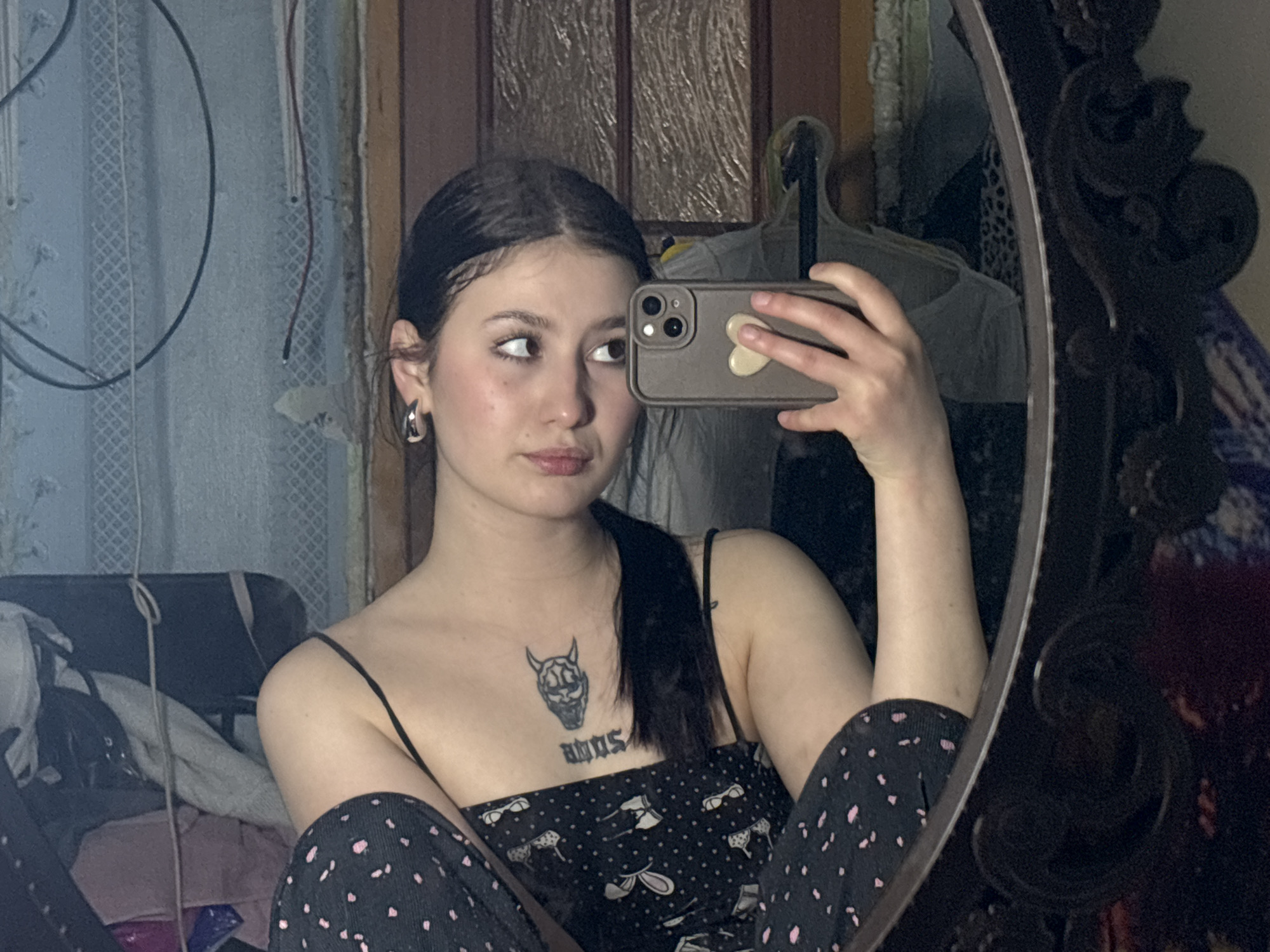 A young woman with dark hair, parted in the middle and pulled back, is taking a selfie in a mirror. She has light skin, dark eyes, and a tattoo on her chest depicting a stylized skull above text. She wears a black tank top with a pattern of cartoon animals and polka-dotted pants. Her left ear has a hoop earring. She holds a smartphone in her right hand, its brown case adorned with a heart-shaped sticker.
The mirror's ornate dark frame is visible on the right side. The background shows a room with a blue patterned wall, a door with glass panels, and clothes hanging on a rack. Clutter is visible in the lower left, including clothes and a dark object resembling a chair.
The location cannot be determined based on the given image alone. The statement that it is in the city of Фоки, Russia cannot be verified.