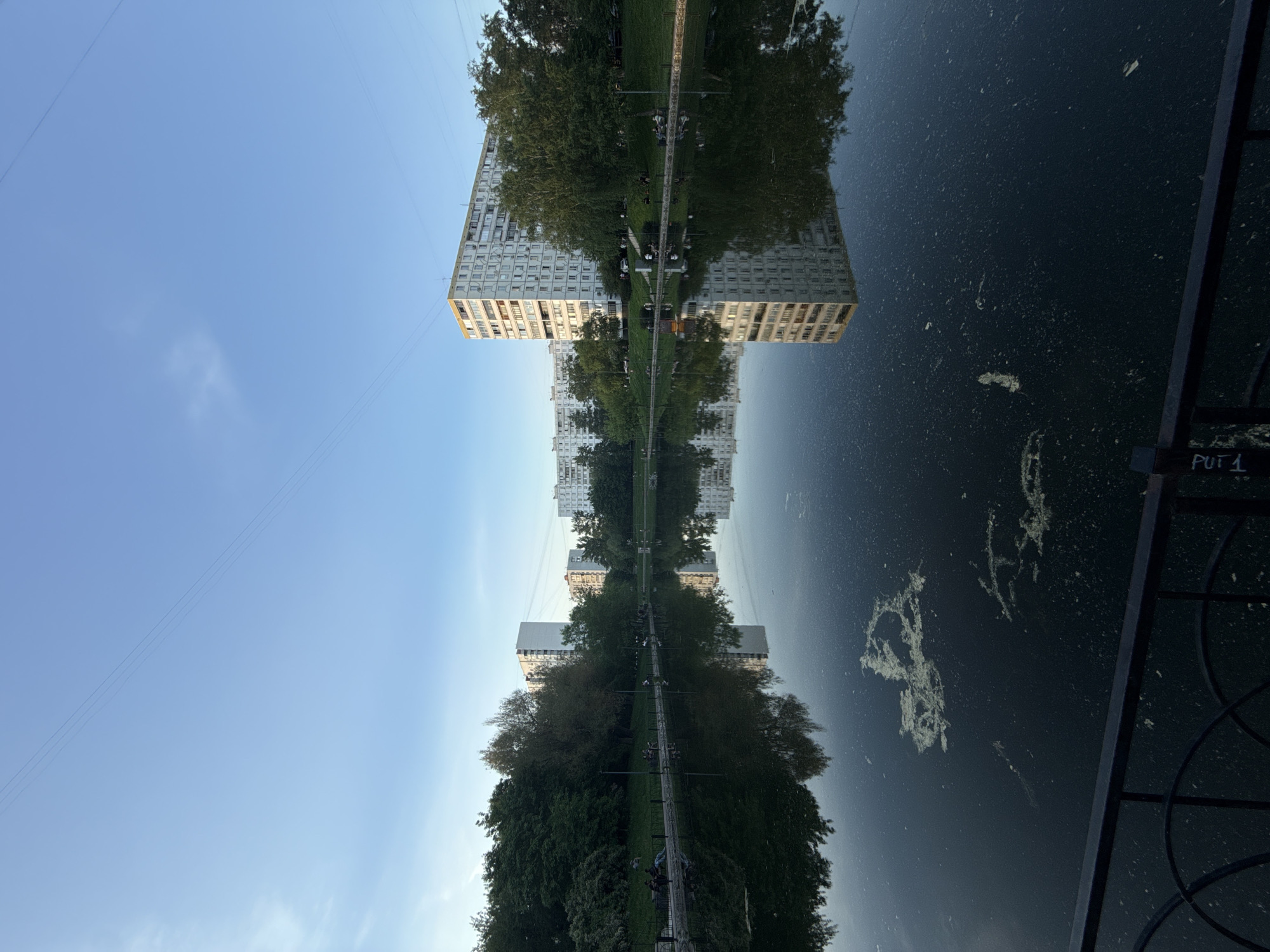 The image shows a reflection in water, likely a pond or lake, of buildings and trees. The sky is visible above the buildings and in the reflection. The image appears to be rotated 180 degrees, as the sky is at the bottom of the image. "PUT 1" is written on an object on the right side of the image. Given the context of being in Moscow, Russia, it is likely that the buildings are typical Soviet-era apartment blocks found in many areas of the city. People and interactions are not clearly visible, though there appear to be people on a pedestrian path in the center of the image. The environment consists of urban structures combined with green spaces.