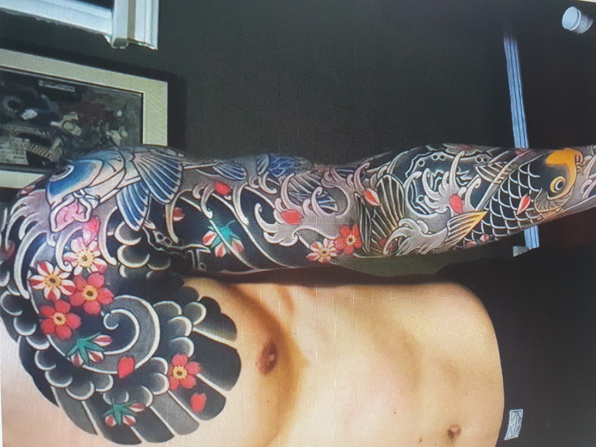 A person is partially visible from the chest down, covered in a large, intricate tattoo. The tattoo covers the left side of their chest and extends down their left arm. It features a traditional Japanese style with elements like koi fish, swirling water, and flowers. The person is wearing black underwear. The background is a dimly lit room with a framed picture hanging on the wall. The overall impression is of a close-up shot focused on the tattoo. There is no indication of the city of Tashkent, Uzbekistan, or any other specific location.
