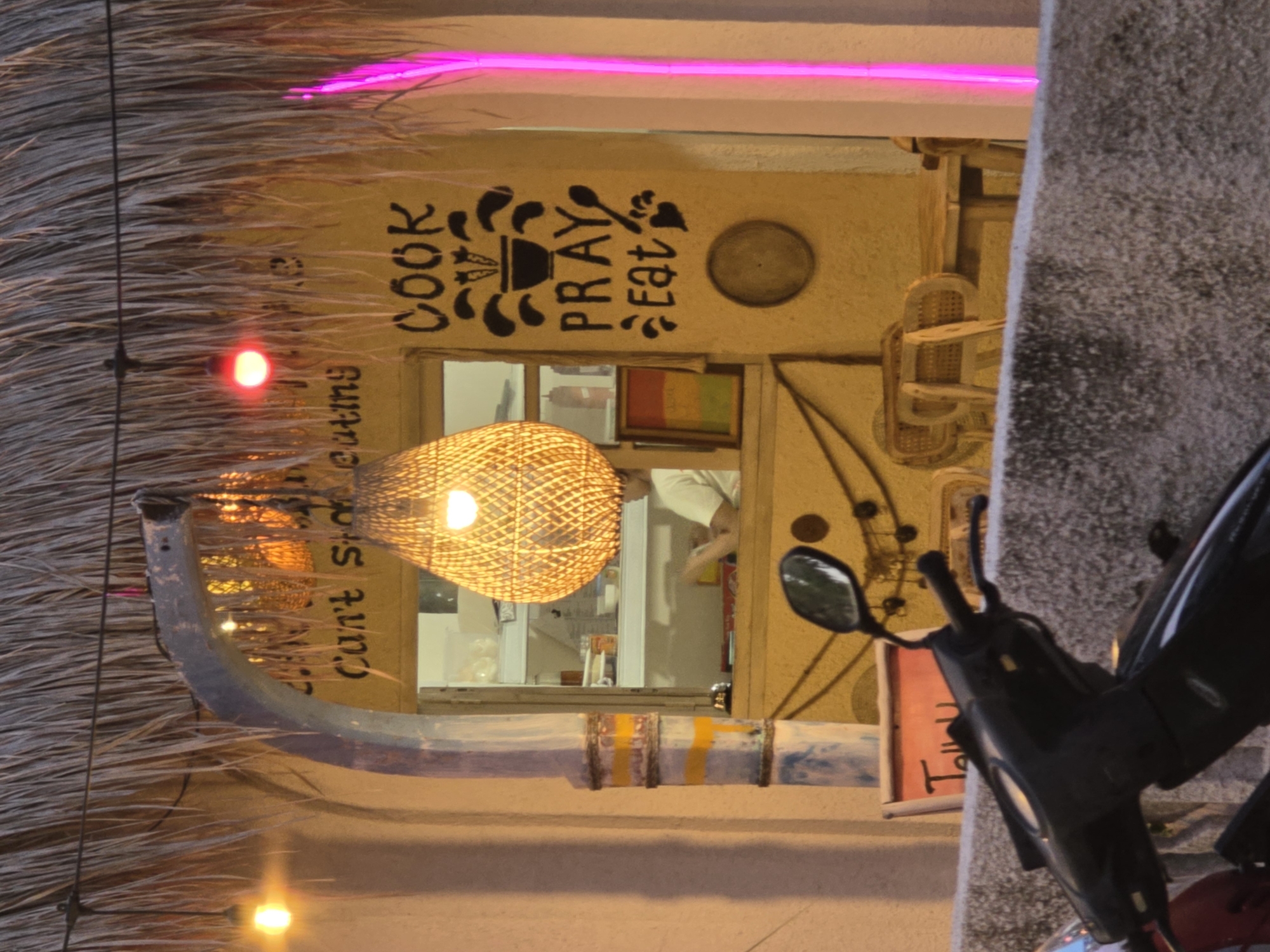 The image shows a building with a thatched roof in an urban environment. A sign reads "Cook Pray Eat". A motorcycle is parked in front of the building. A woven light fixture hangs in front of a window. The location of the image is Unknown, Unknown. The motorcycle is black and red. A neon light can be seen in the upper part of the image. A person is seen inside the building.