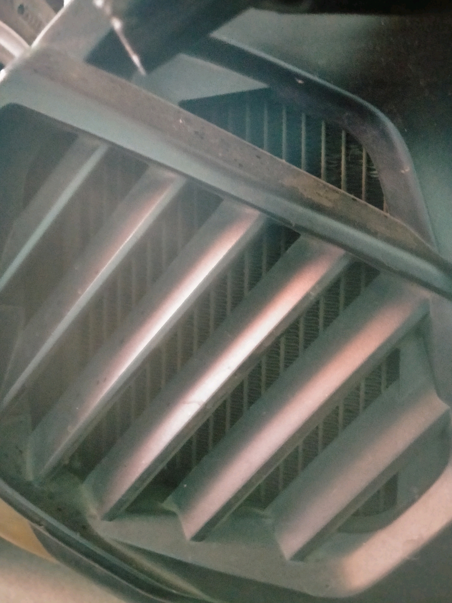 The image shows a close-up, low-angle view of a vehicle's front grille and radiator. The grille features prominent horizontal slats with a metallic sheen, interspersed with a darker mesh-like material behind it, likely part of the radiator fins. The lighting suggests it is outdoors, possibly in daylight, with shadows and highlights emphasizing the contours of the grille. The scene appears to be a static shot, with no people or active elements present. The location, Klepu, Indonesia, is a village in Central Java, known for its agricultural landscape and rural setting. While the image itself doesn't offer direct clues to this specific location, it depicts a common automotive component found throughout Indonesia.