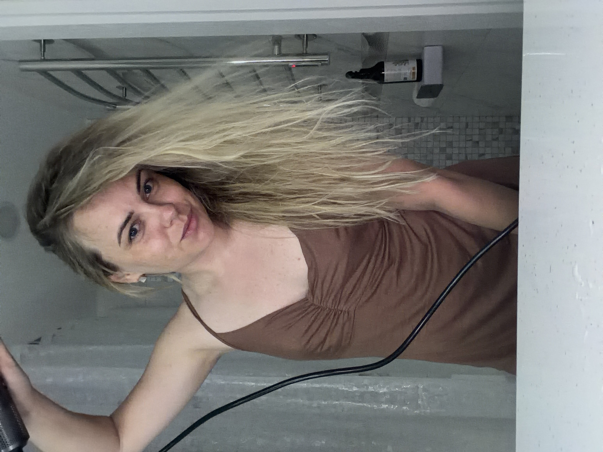 A woman with blonde hair and blue eyes is in a bathroom. She's wearing a brown dress with thin straps. She's holding a black hair dryer. There is a shower head, towel rack, bottle and white dispenser on the wall behind her. The bathroom has mosaic tiles. The location is likely not Votkinsk, Russia as there is no evidence to support that claim.