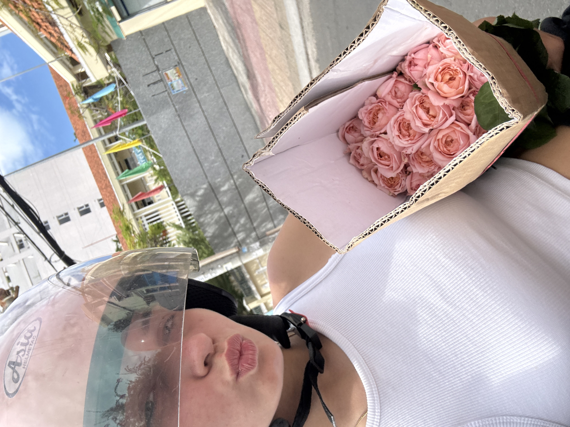 A woman wearing a pink helmet with a clear visor and a white tank top is holding a cardboard box filled with pink roses. She is on a scooter, as indicated by the helmet and her position. The background shows a street scene with buildings, flags, and power lines, suggesting an urban environment. The woman is making a kissing gesture towards the camera. The scene appears to be in Nha Trang, Vietnam, based on the visual cues of the architecture and the tropical foliage.