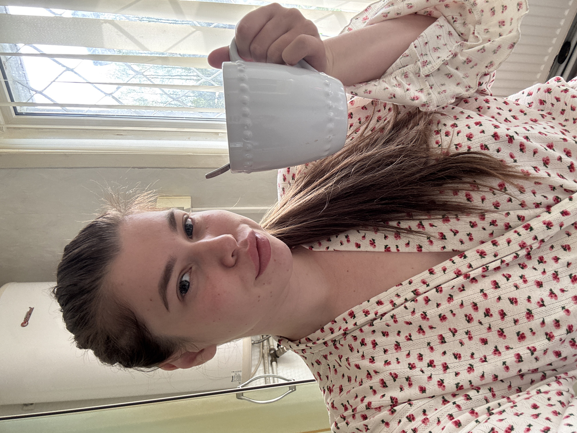 A young woman is holding a white mug with a spoon in it. She is wearing a white top with a red floral pattern. The background shows a window with blinds, a kitchen counter, and a glimpse of kitchen fixtures. The location is not identifiable as Мещёры, Russia from this image.