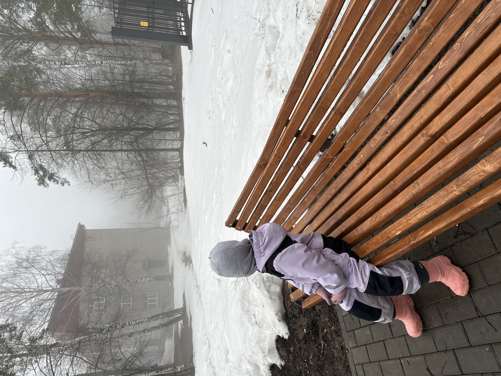 A young child is lying on a wooden park bench outdoors. The child is wearing a grey hat, a lavender jacket, black pants, and pink boots. The background shows a snowy area with trees and a building obscured by fog. The scene is likely located in Chaikovsky, Russia, given the request to identify the location. The ground around the bench is a combination of snow and dark soil. The weather appears cold and overcast. The child is the primary subject and is the only person visible in the image. The child is lying down, suggesting rest or relaxation. The environment appears quiet due to the fog. The surface under the bench is made of gray bricks.