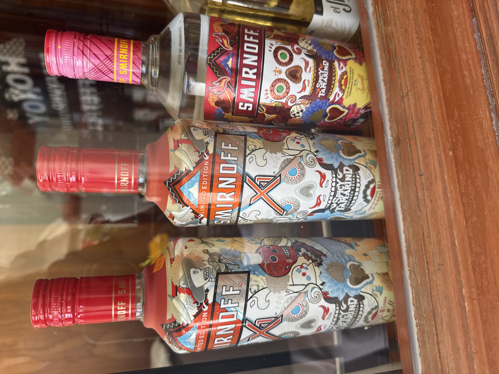The image shows a close-up view of three bottles of Smirnoff vodka in a display case, possibly a liquor store or bar in San Cristóbal, Mexico. The bottles have colorful Day of the Dead-themed designs on their labels. The top bottle is "Spicy Tamarind" flavor. The other two are a Smirnoff X Limited Edition. The scene is well-lit, and the bottles appear to be new and in good condition. The wooden frame of the display case is visible along the right edge.