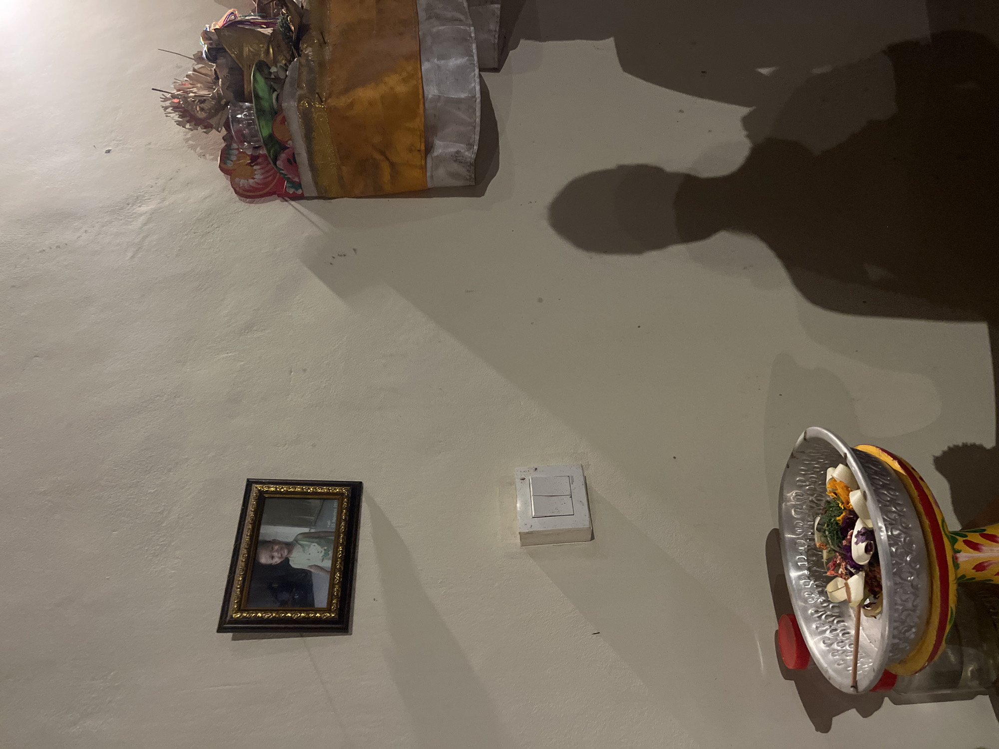 A dimly lit indoor scene in Kuta Selatan, Indonesia, reveals a textured, light-colored wall serving as the backdrop for a collection of objects. At the top left, a decorative arrangement features fabric and floral elements. Moving downward, a framed portrait with a gold-patterned frame hangs on the wall. To the right, a light switch plate is mounted, and further to the right, a circular tray filled with various items is affixed to the wall. A shadow of a person is cast on the wall.