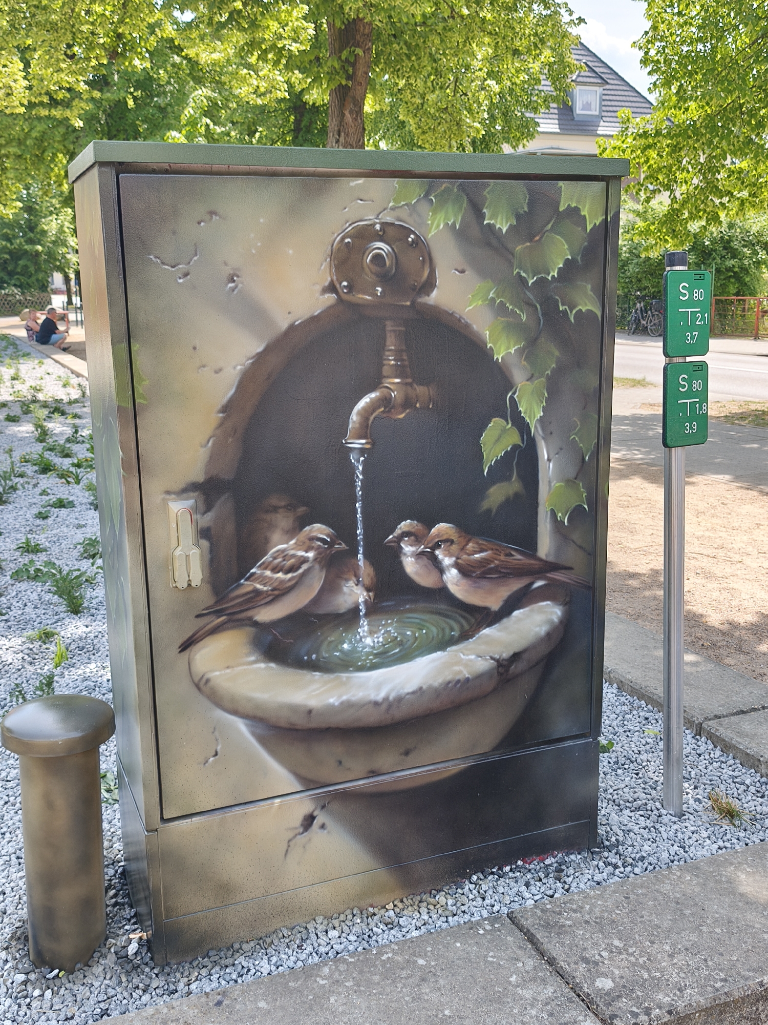 The image depicts a utility box in an outdoor setting, possibly in Unknown, Unknown. The box is painted with a detailed mural of a stone birdbath with a tap, water, and several sparrows. To the left of the box is a small, cylindrical metal object. To the right, a metal pole holds two green signs, each marked with "S 80" along with other numbers. The box and pole are surrounded by small white stones. Beyond the box, there is a paved area, grass, trees, and in the distance, a building with a gray roof. A few people can be seen sitting in the background on the left.