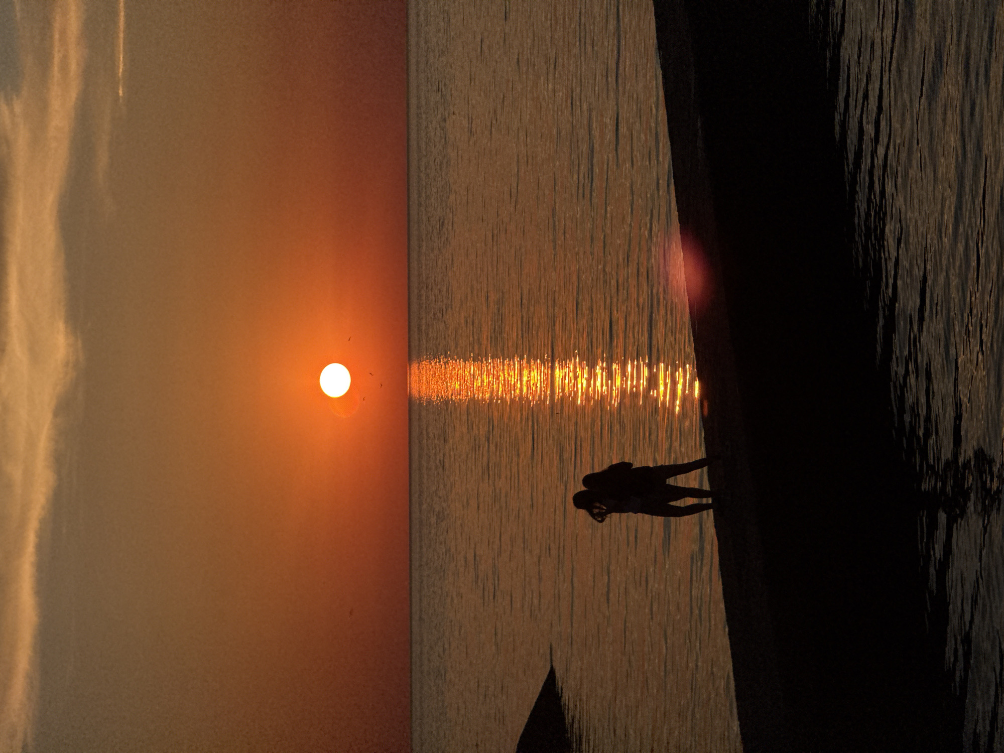 A vertical, eye-level, full shot captures the setting sun over a tranquil ocean. The sun, a bright white orb, is centered in the frame, casting a shimmering golden path across the water. Two figures, silhouetted against the warm orange sky, stand on a dark, sandy shore in the foreground. They are positioned side-by-side, with one figure appearing to hold a camera and point it towards the horizon, possibly taking a photograph of the sunset. The water is a deep, muted orange, reflecting the sun's glow. The sky transitions from a fiery orange at the horizon to a softer, dusky hue higher up. Wispy clouds drift lazily across the sky. A dark, rocky outcrop is visible on the left side of the frame, partially submerged in the water. The overall impression is one of peaceful serenity and natural beauty.