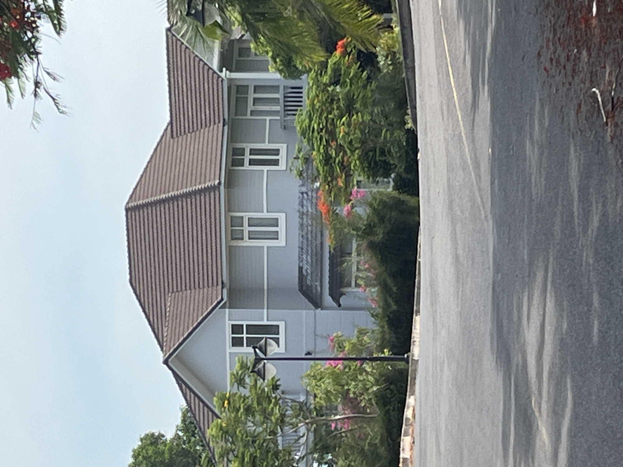 The image shows a two-story house with a gray exterior and a brown tile roof. The house has multiple windows with white frames. In front of the house, there are trees and flowering bushes, some with pink and orange blossoms. A black lamppost stands in the foreground, to the left of the house. The house appears to be situated in a residential area, possibly in the city of Phu Thuy Ward, Vietnam, given the lush vegetation and architectural style. The foreground is dominated by a paved road, with some shadows cast on it from the trees.