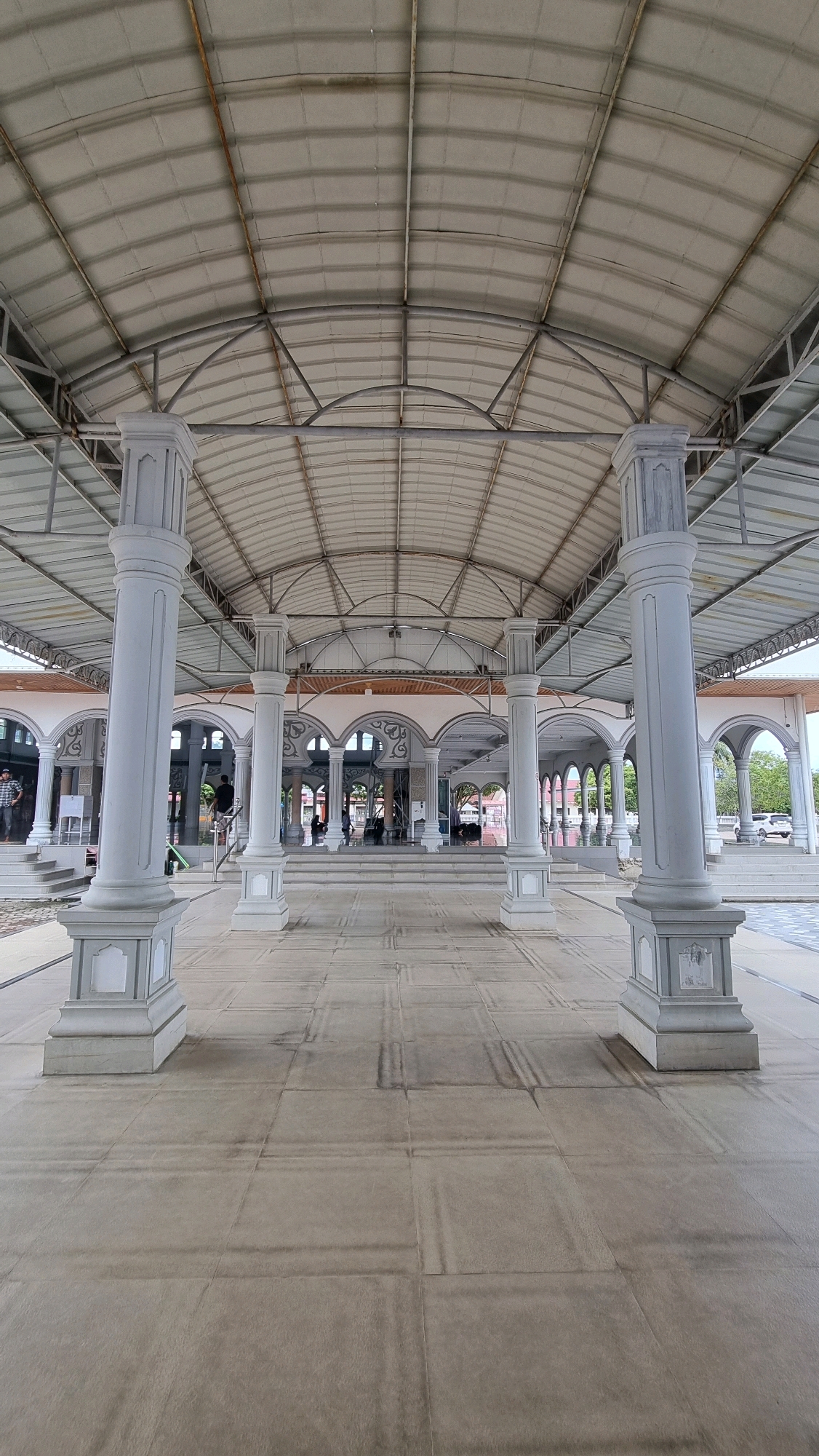 The image depicts the exterior walkway of a mosque in Manetunong, Indonesia. The walkway is covered by a large, arched, metal roof supported by rows of white, fluted columns. The floor is paved with large, light-colored tiles arranged in a grid pattern, with some darker, linear markings visible on the surface. In the background, there are more arched structures and entrances leading into the mosque. Several people are visible, some standing and some walking, but their faces are not clearly discernible. The overall scene suggests a public, religious space with an open-air, covered area.