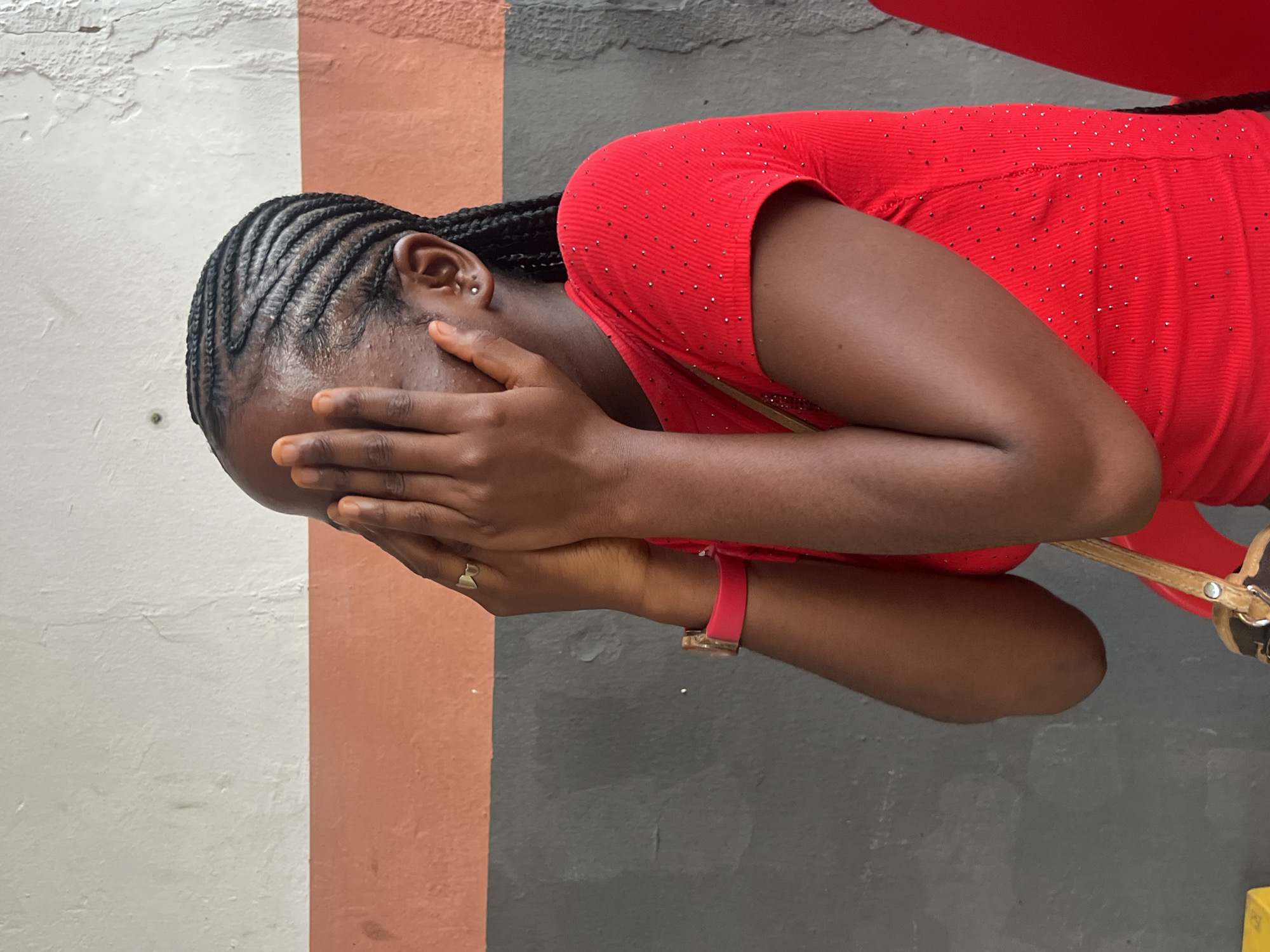 The image shows a young woman with braided hair covering her face with her hands. She is wearing a red, sparkly shirt and a pink watch on her left wrist. A ring is visible on her right hand. She appears to be standing against a wall painted in three horizontal sections: white at the top, orange in the middle, and dark gray at the bottom. A portion of a brown and black patterned bag strap is visible on her left shoulder. The background wall suggests an outdoor or semi-outdoor setting, possibly in the city of Nkpor, Nigeria, given the context.