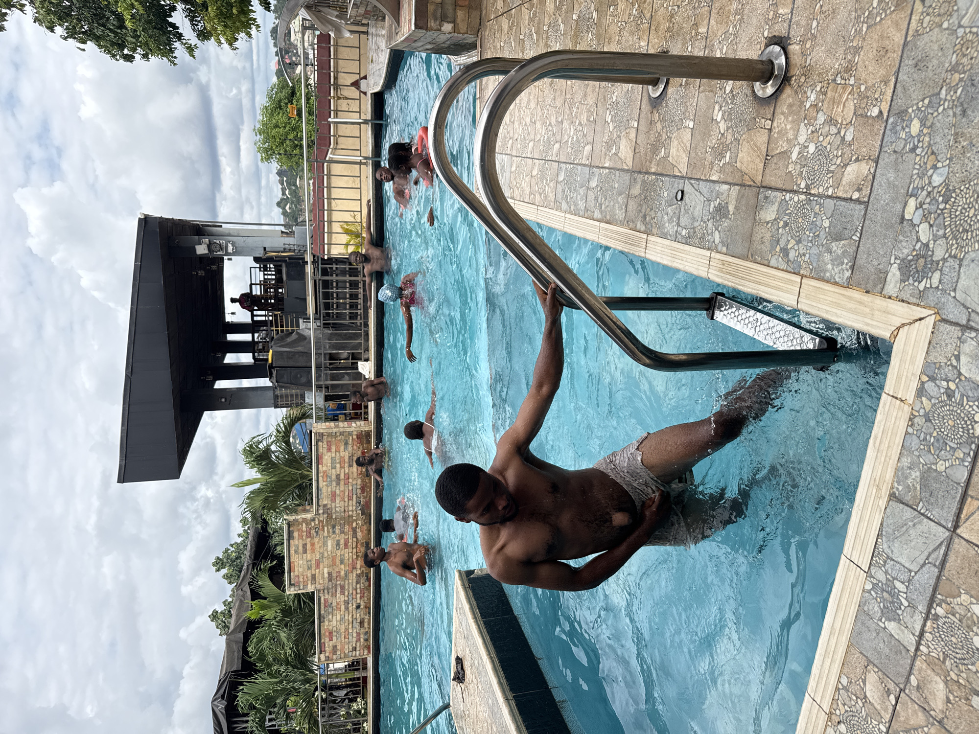 A group of young Black men and at least one woman are swimming and socializing in a clear blue outdoor swimming pool at a resort in Nkpor, Nigeria. The water is calm and the sky is cloudy. A modern building with a black roof is visible in the background, and there are palm trees and other greenery surrounding the pool area. The man in the foreground is exiting the pool, holding onto the stainless steel railing of the steps, wearing patterned swim shorts. Other individuals are spread throughout the pool, some appearing to be conversing, while others are swimming or relaxing in the water. The pool deck is tiled with a mosaic pattern.