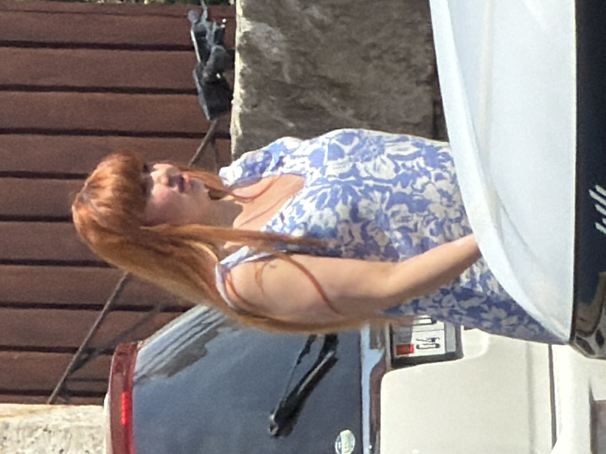 A woman with long, auburn hair stands outdoors, facing left, wearing a blue and white floral dress. She is positioned to the right of a white car with a red brake light and a partial license plate visible. The background includes a textured brown wall and a stone wall. The lighting suggests it is daytime, casting shadows.