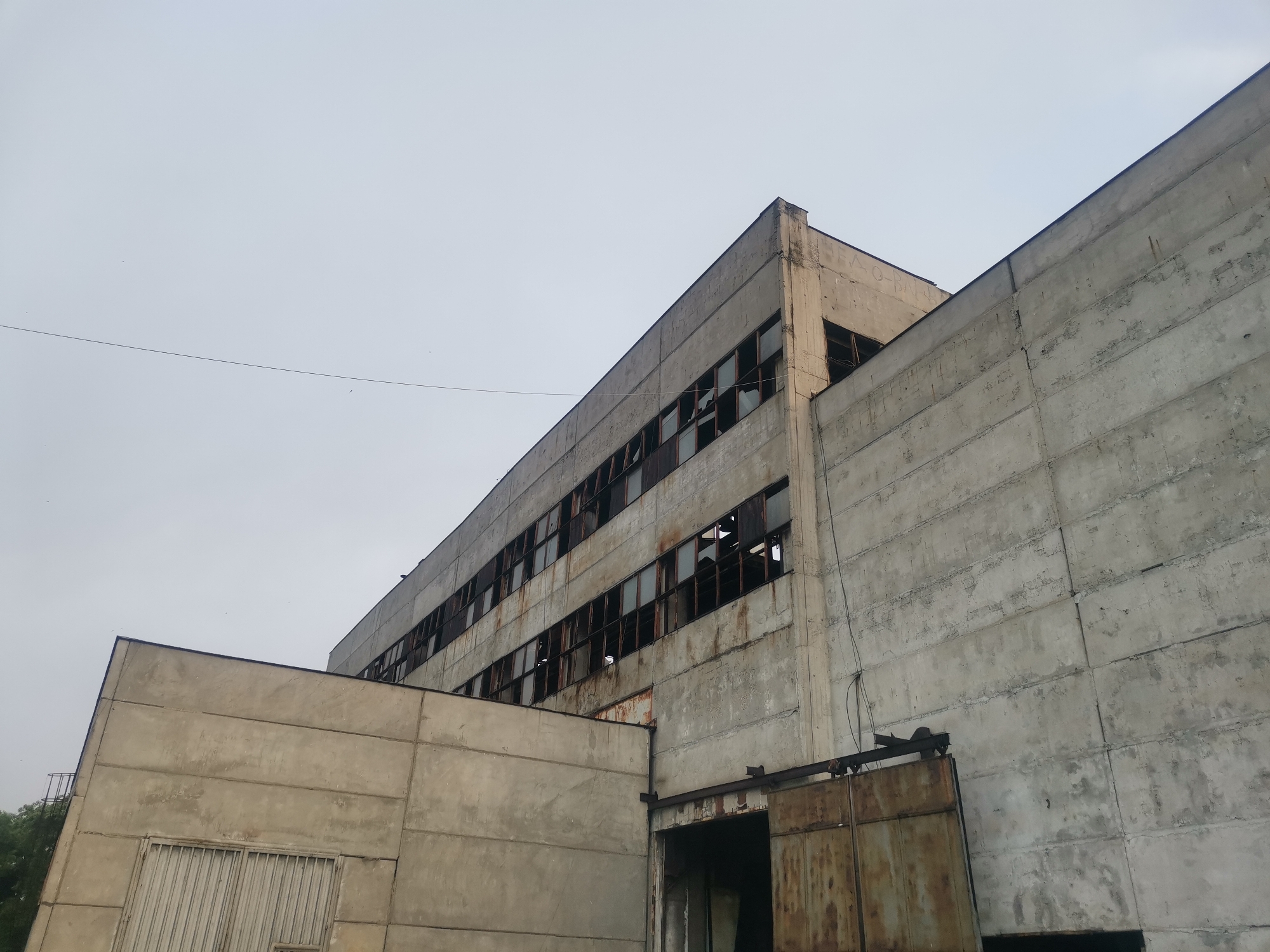 This image presents an exterior view of a large, disused industrial building in Zhivkovo, Bulgaria, under an overcast sky. The structure is predominantly made of rough, weathered concrete panels, displaying discoloration, dirt, and streaking. The main section of the building features multiple rows of windows with broken panes and rusted metal frames, offering glimpses into dark, unlit interiors.
At ground level, a large, heavily rusted metal sliding door or gate is partially open, revealing a dark entrance. To the left, a smaller, lower concrete annex with a sloped roof abuts the main structure, featuring a metal grate or barrier on its wall. A thin overhead wire traverses the upper portion of the frame against the grey sky.
The scene depicts a static, abandoned environment. No people are visible, and no active events are taking place. The overall impression is one of decay and neglect. Faint Cyrillic graffiti, appearing to read "ТРАОРН" (meaning "mourning" or "grief"), is discernible on the upper concrete wall near the roofline, adding to the somber atmosphere. The time of day is clearly daylight, but the lack of direct sunlight indicates cloudy weather.
