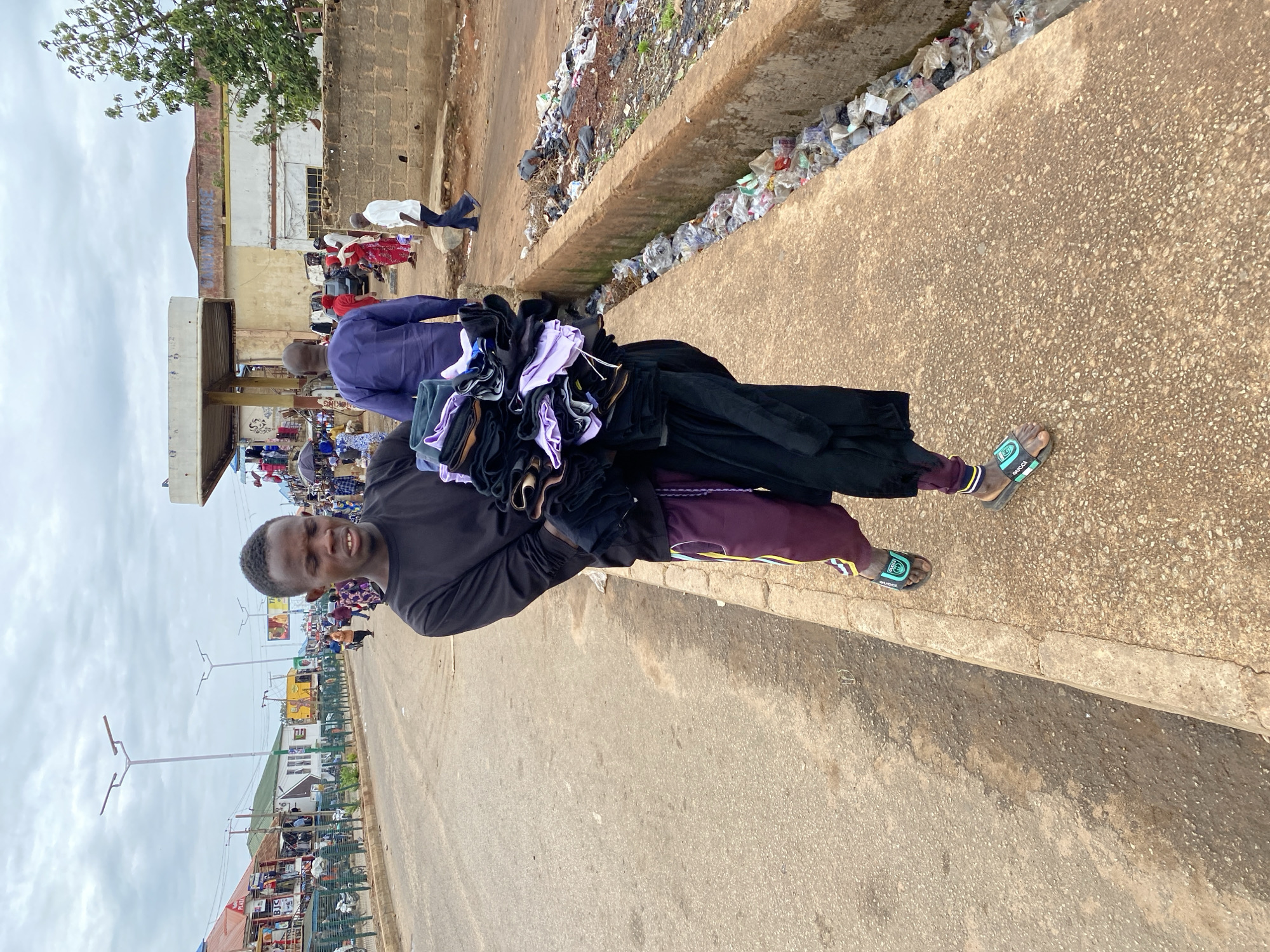 In Bukuru, Nigeria, a young Black male is captured mid-stride on a concrete sidewalk, his gaze directed forward with a slightly strained expression. He is carrying a large bundle of folded clothing, predominantly black and purple, held close to his chest. His attire consists of a long-sleeved black shirt and maroon pants adorned with yellow and white stripes along the sides, complemented by designer-style slides on his feet. The background reveals a bustling street scene: a paved road with vehicular and pedestrian traffic, shops lining the left side, and a prominent overpass structure with shops beneath it. To the right of the sidewalk, a narrow, open concrete drain is visible, filled with accumulated trash, including plastic bottles and bags, alongside scattered debris. The sky is overcast, suggesting a cloudy day. The overall environment depicts a commercial area in an urban setting.