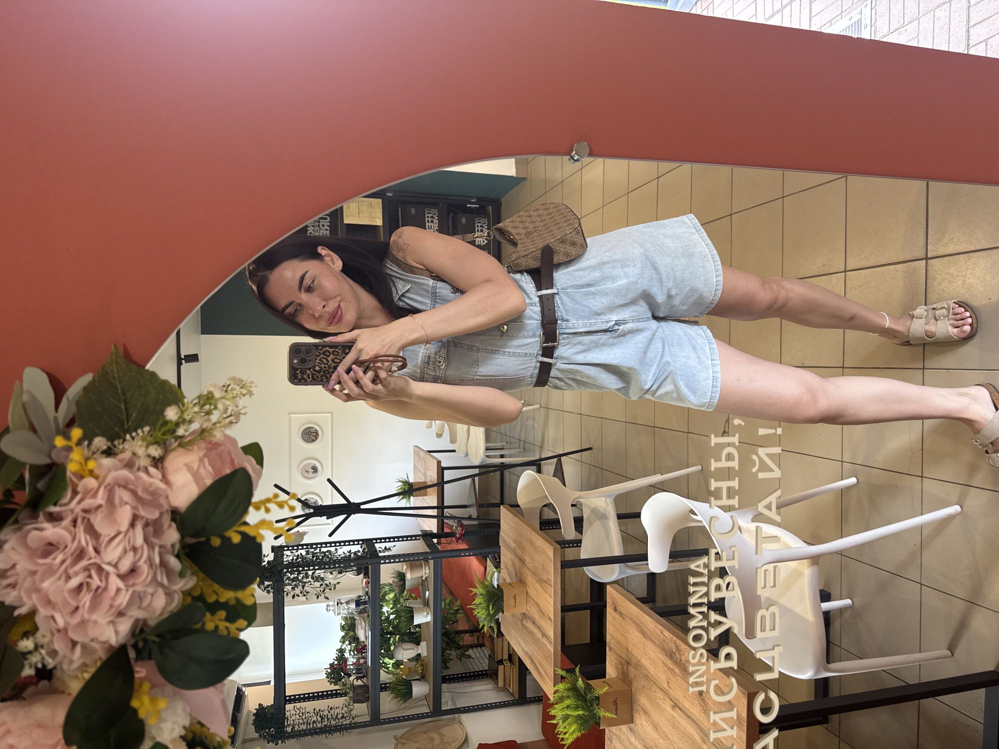 The image captures a woman taking a mirror selfie in what appears to be a cafe or restaurant. The setting includes light-colored tiled flooring, wooden tables with metal legs, and white chairs. In the background, shelves with plants and decor are visible. A prominent floral arrangement hangs from the top of the mirror. The woman is wearing a light blue denim romper, a brown belt, and beige sandals. She is holding a smartphone with a leopard-print case, taking the photo. Text is overlaid on the lower part of the mirror, reading "INSOMNIA" and then Cyrillic text. The overall scene suggests a casual, indoor environment. There is no visual information in the image that would place the location specifically in Chaikovsky, Russia.