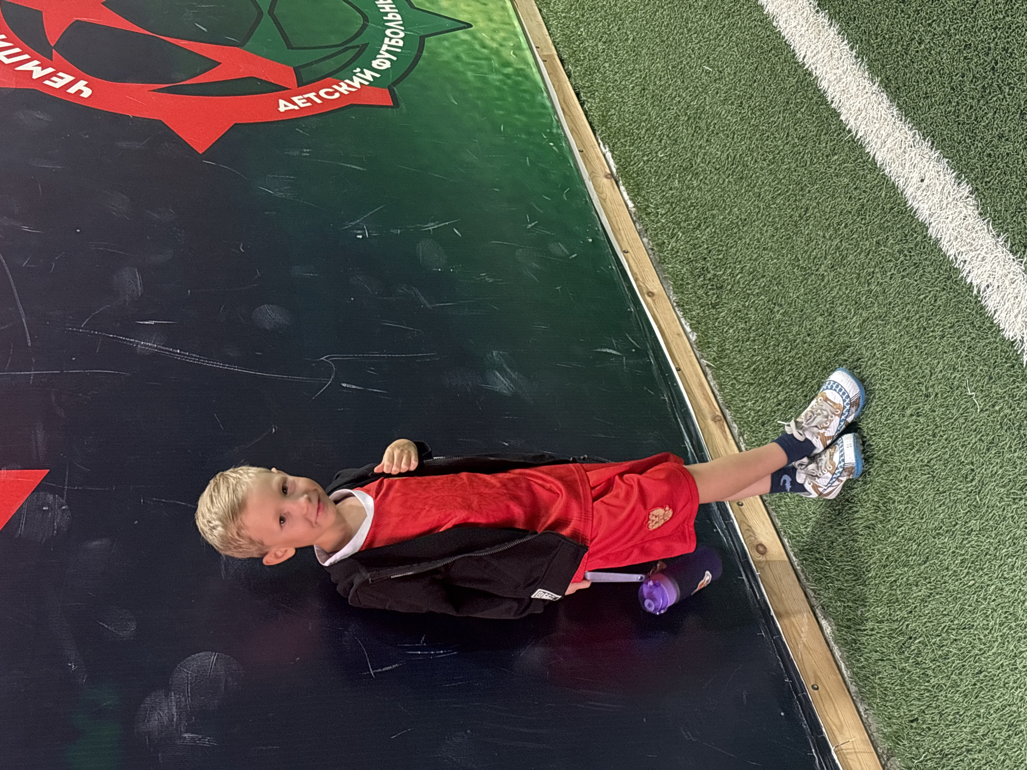 A young boy with blonde hair is standing against a dark, textured wall. He is wearing a red t-shirt, red shorts with an emblem on the left leg, a black zip-up jacket, dark socks, and white and blue sneakers. He is holding a purple water bottle with a black lid in his left hand. The wall behind him has green and red graphics. The floor in front of him is green artificial turf with a white line marking. The scene appears to be indoors, possibly at a sports facility, given the turf and the graphics on the wall, which include text in Cyrillic that may indicate a championship or sports event. The lighting is somewhat dim, casting shadows. The boy is looking towards the camera with a slight smile. The overall impression is of a child at a sports-related event.