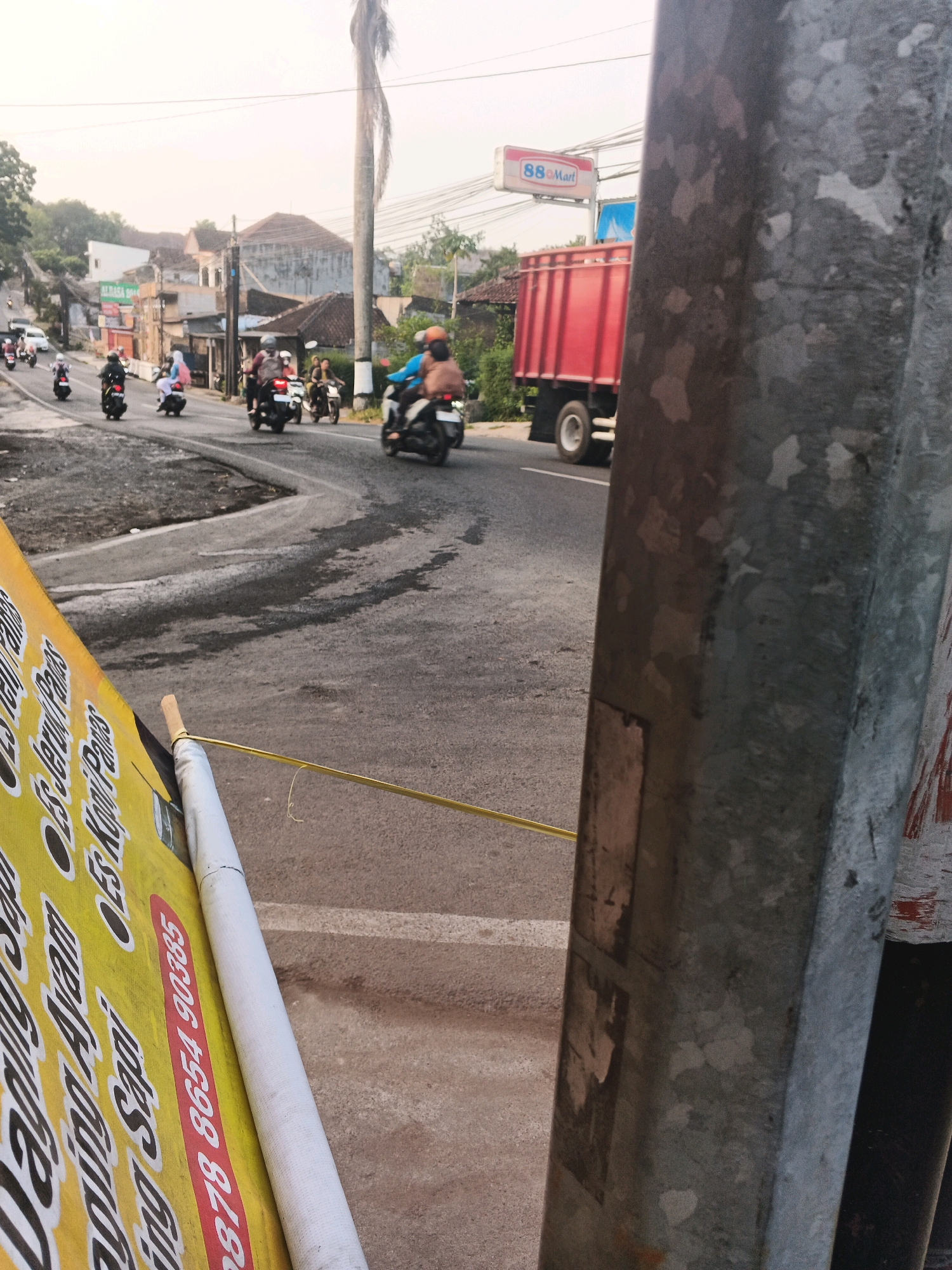 This is a street-level shot, likely in Semarang, Indonesia, capturing a busy road scene. In the foreground, a yellow banner with black text is partially visible, angled to the left. The text on the banner appears to be an advertisement, with some words like "Sapi", "Ayam", "Jeruk", and "Kopi" discernible, along with a phone number "0878 8654 90385". To the right, a rough, weathered metal pole dominates the frame, showing patches of wear and tear.
The middle ground shows a road with several motorcycles in motion, some carrying one or two riders. The riders are wearing helmets and casual clothing. A large red truck with a high side panel is driving away from the viewer on the right side of the road. In the background, there are buildings, including one with a prominent red sign that says "88 Mart". Power lines crisscross overhead. The sky is overcast and the lighting suggests it might be late afternoon. The overall impression is of a typical, active urban environment with everyday traffic.